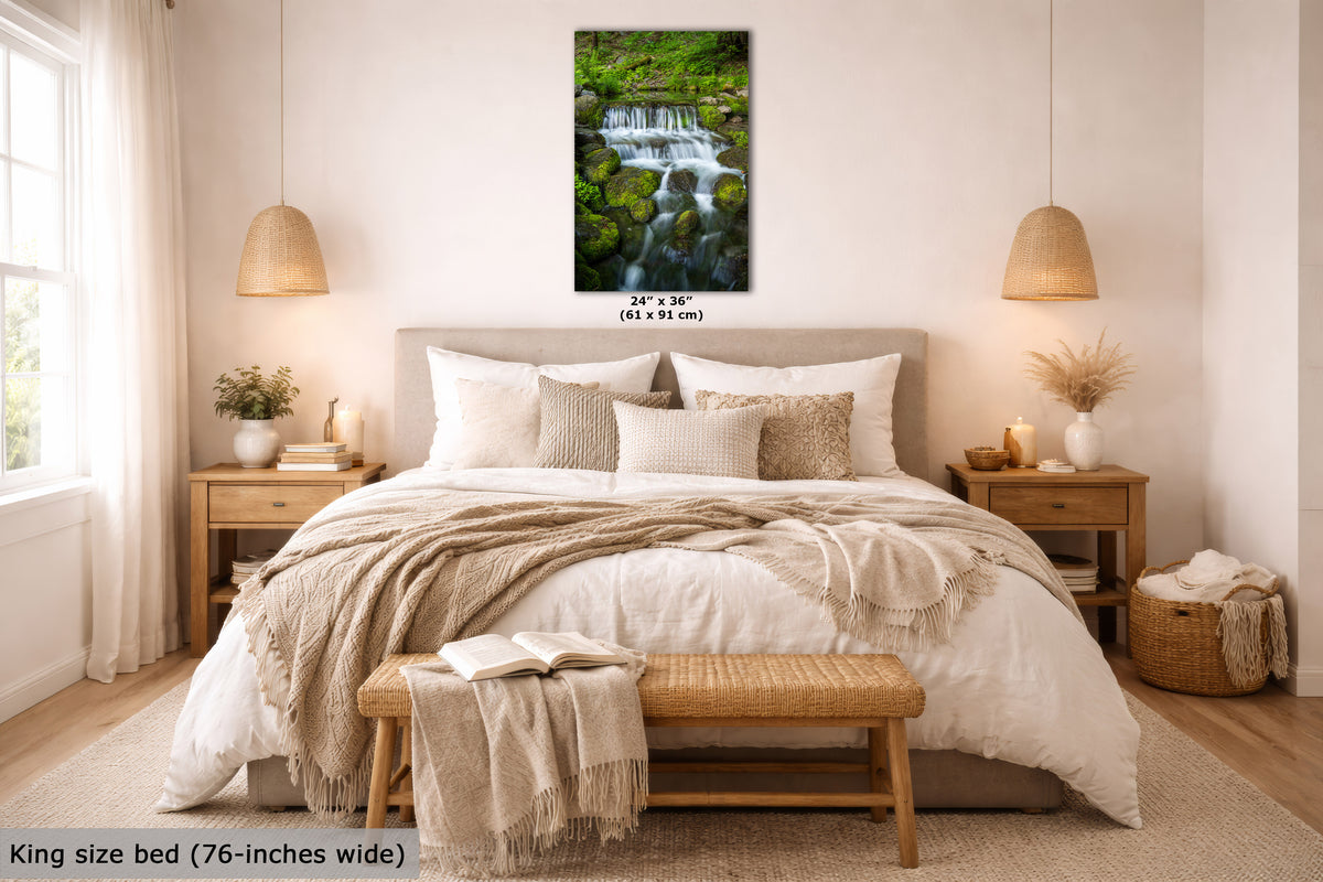 A framed vertical wall art print showing a lush green waterfall landscape, displayed above a neatly made bed in a bright, modern bedroom. The print includes size text indicating 24 x 36 inches (61 x 91 cm).