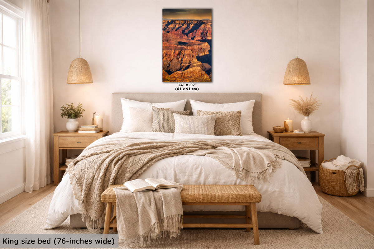 A framed wall art print depicting a canyon landscape in warm orange, brown, and black tones, displayed above a neatly made bed in a bright, modern bedroom. The print includes size text reading 24 x 36 inches, and the room scene includes a bed, nightstands, lamps, and decorative textiles.