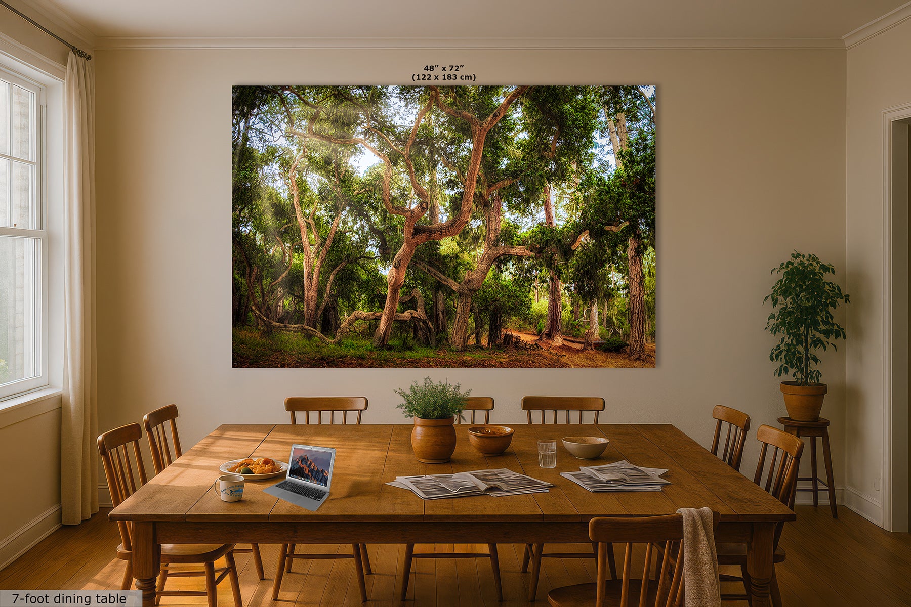 A large rectangular wall art print depicting a sunlit coast live oak forest with tall trees and green foliage, displayed above a wooden dining table in a real dining room with chairs, plants, and windows.