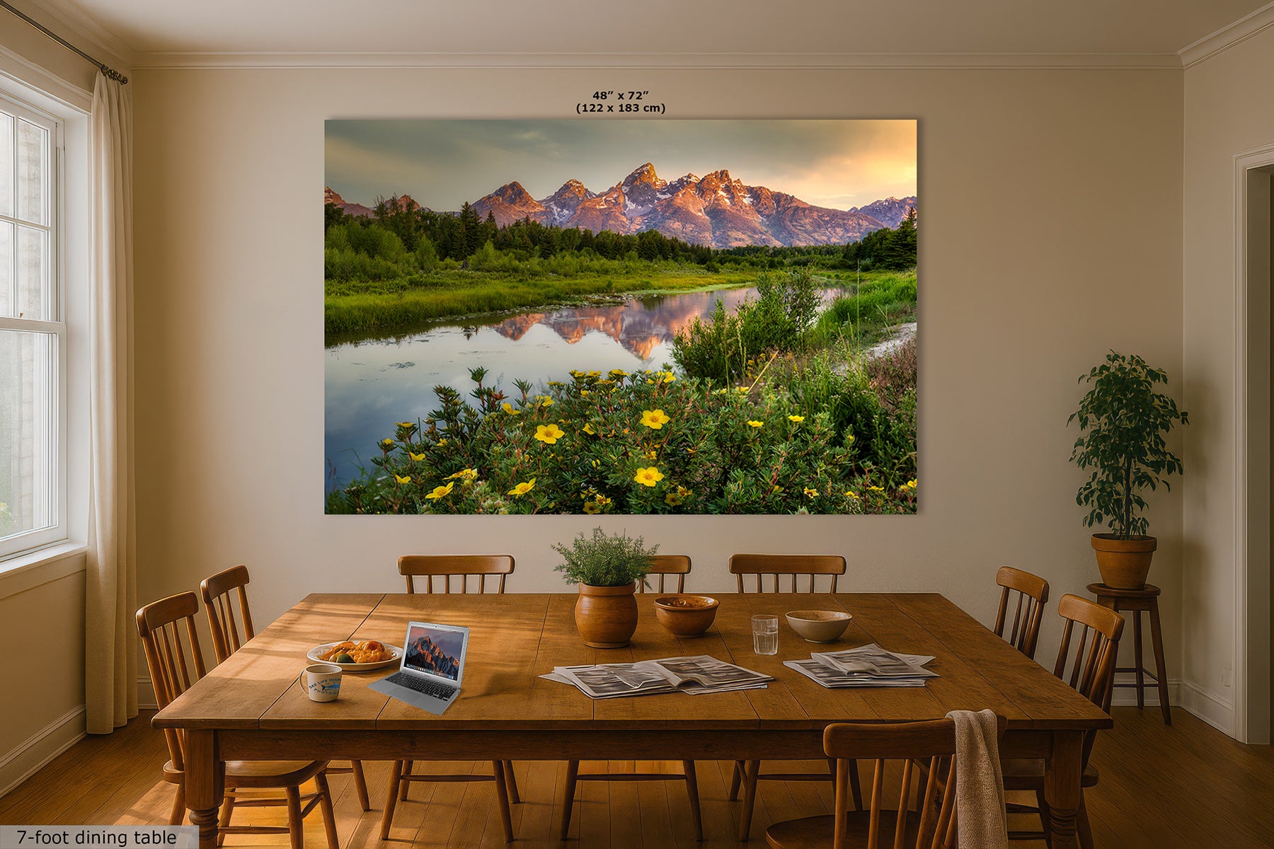 A large wall art print depicting a Grand Teton mountain river landscape with green fields and yellow flowers, displayed above a wooden dining table in a bright dining room. The print includes size text at the top and a small label at the bottom left.