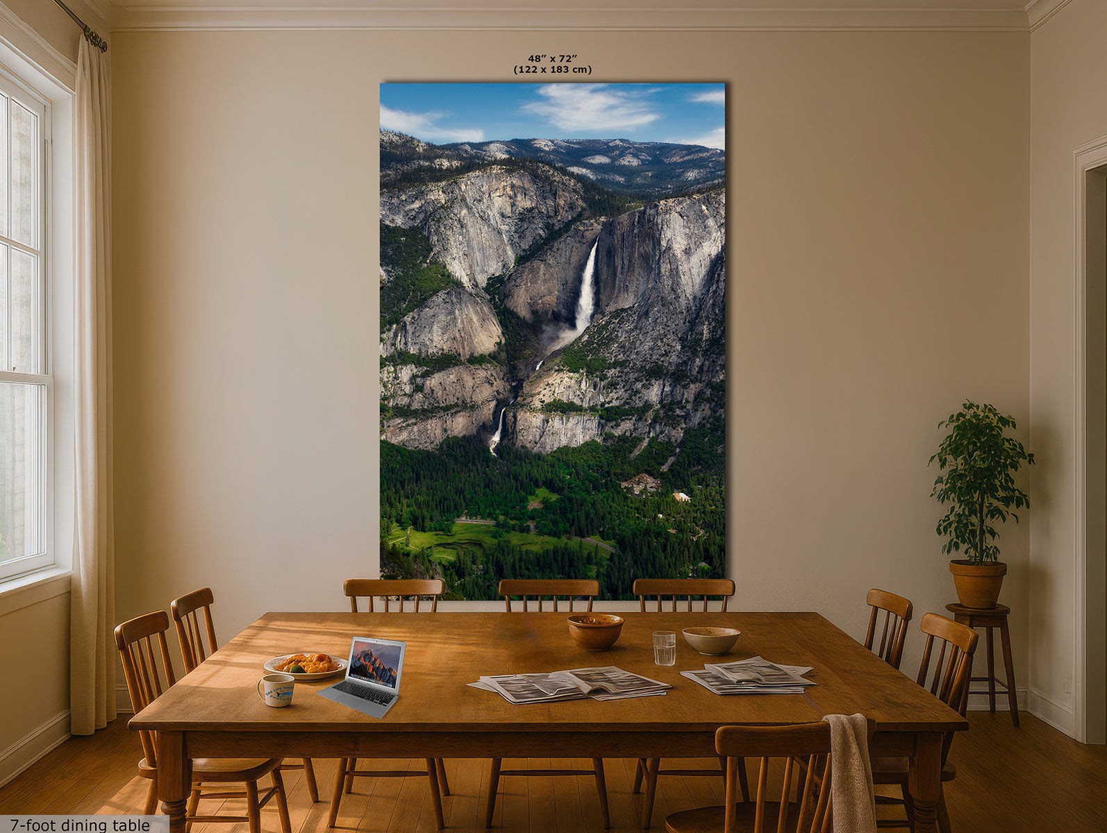 A large vertical wall art print depicting a mountain landscape with a waterfall, displayed above a wooden dining table in a beige dining room. The print includes size text at the top and a small '7-foot dining table' label in the lower left corner.