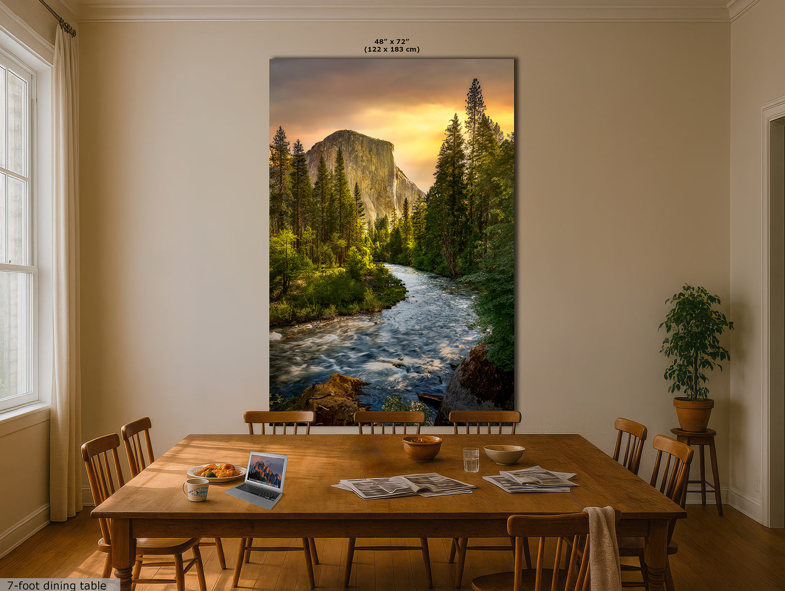 A large vertical Yosemite wall art print of El Capitain and the Merced River that hangs on a beige wall above a wooden dining table in a dining room. The artwork shows a rushing river, dense evergreen trees, and a glowing sunset sky. Small text at the top reads 48 x 72, and a small label at bottom left says 7-foot dining table.