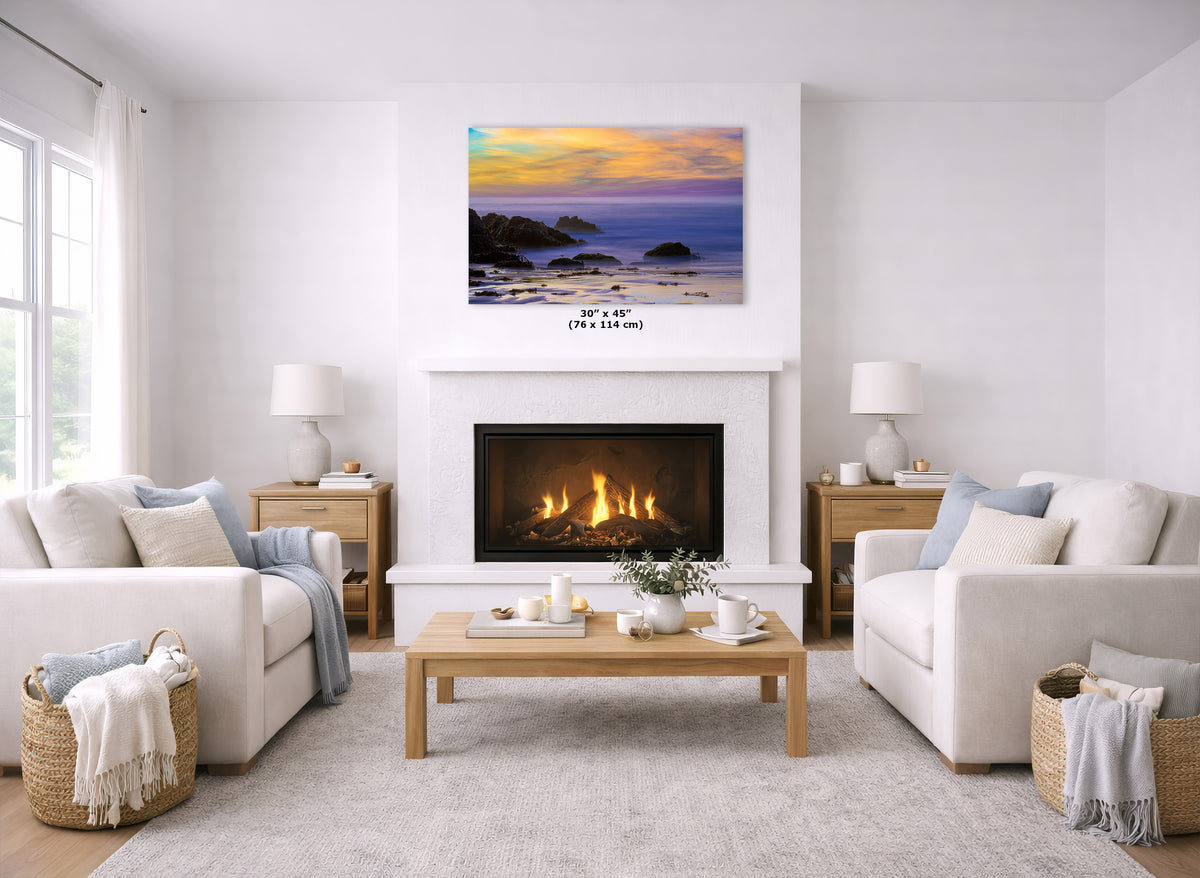A framed coastal sunset wall art print is displayed above a white fireplace in a bright, modern living room with two cream sofas, a wooden coffee table, and a light rug. The print shows a beach and ocean at sunset with blue, purple, orange, and yellow tones.