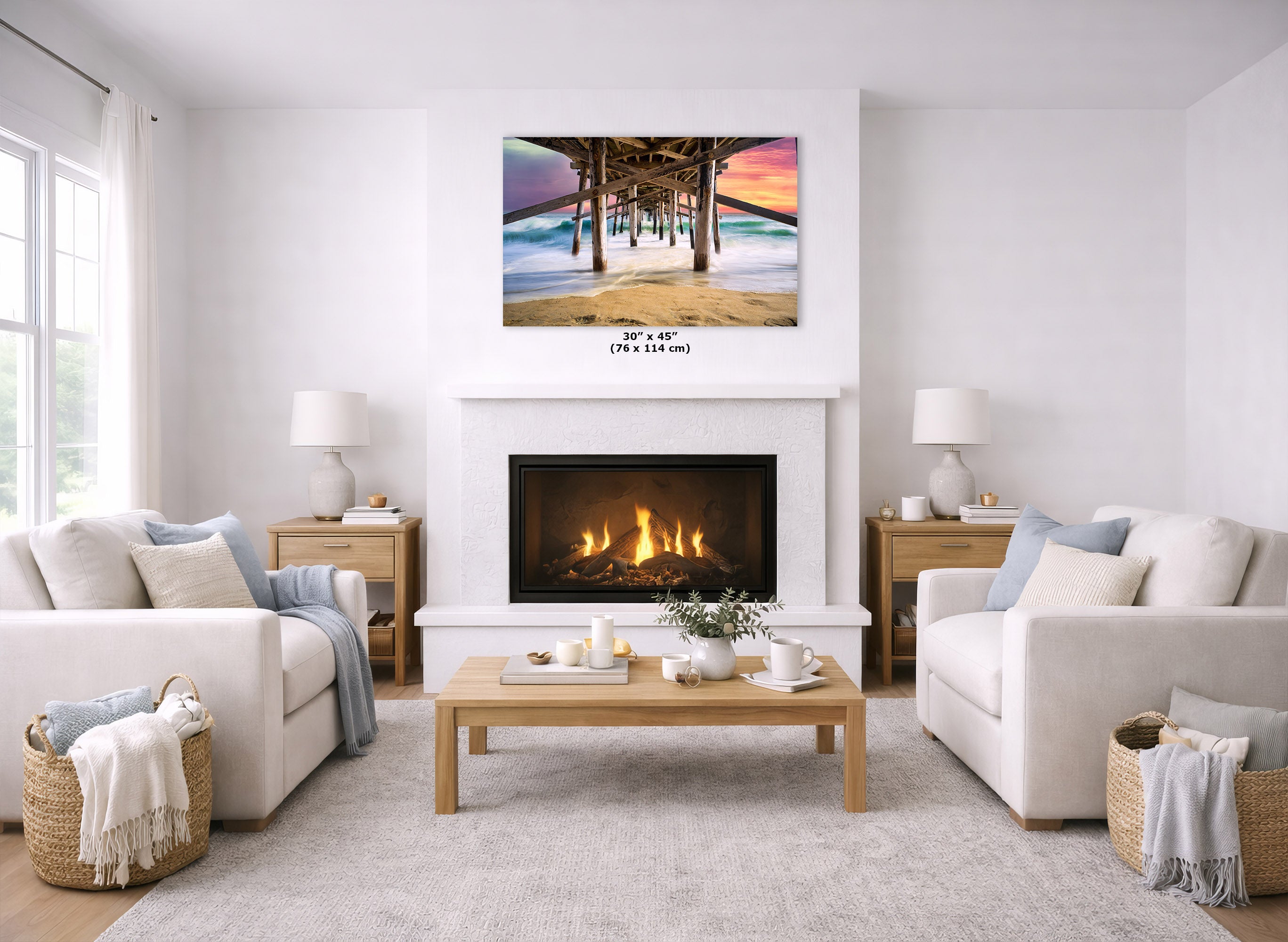 A wall art print depicting a pier over a beach at sunset, displayed above a fireplace in a bright, modern living room with two white sofas, a wooden coffee table, and a light rug.