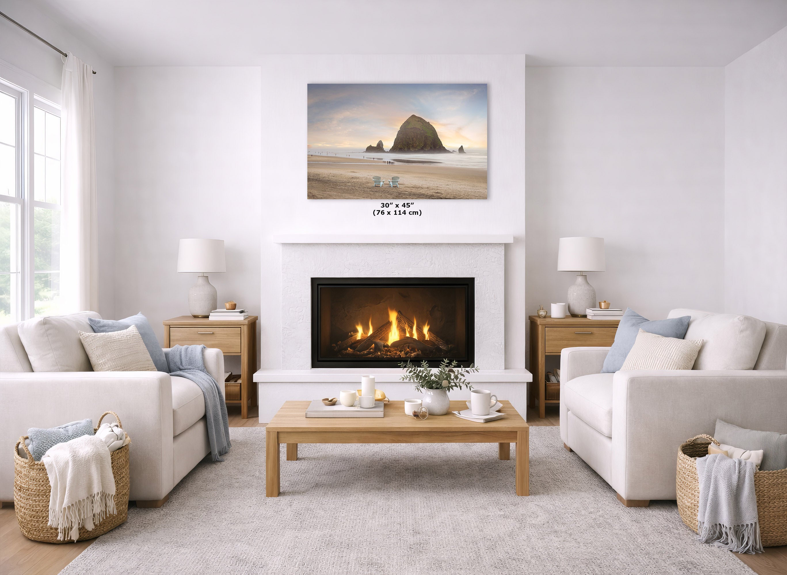 A coastal landscape print of Haystack Rock on a beach is displayed above a white fireplace in a bright, modern living room with two cream sofas, a wooden coffee table, and a light rug.