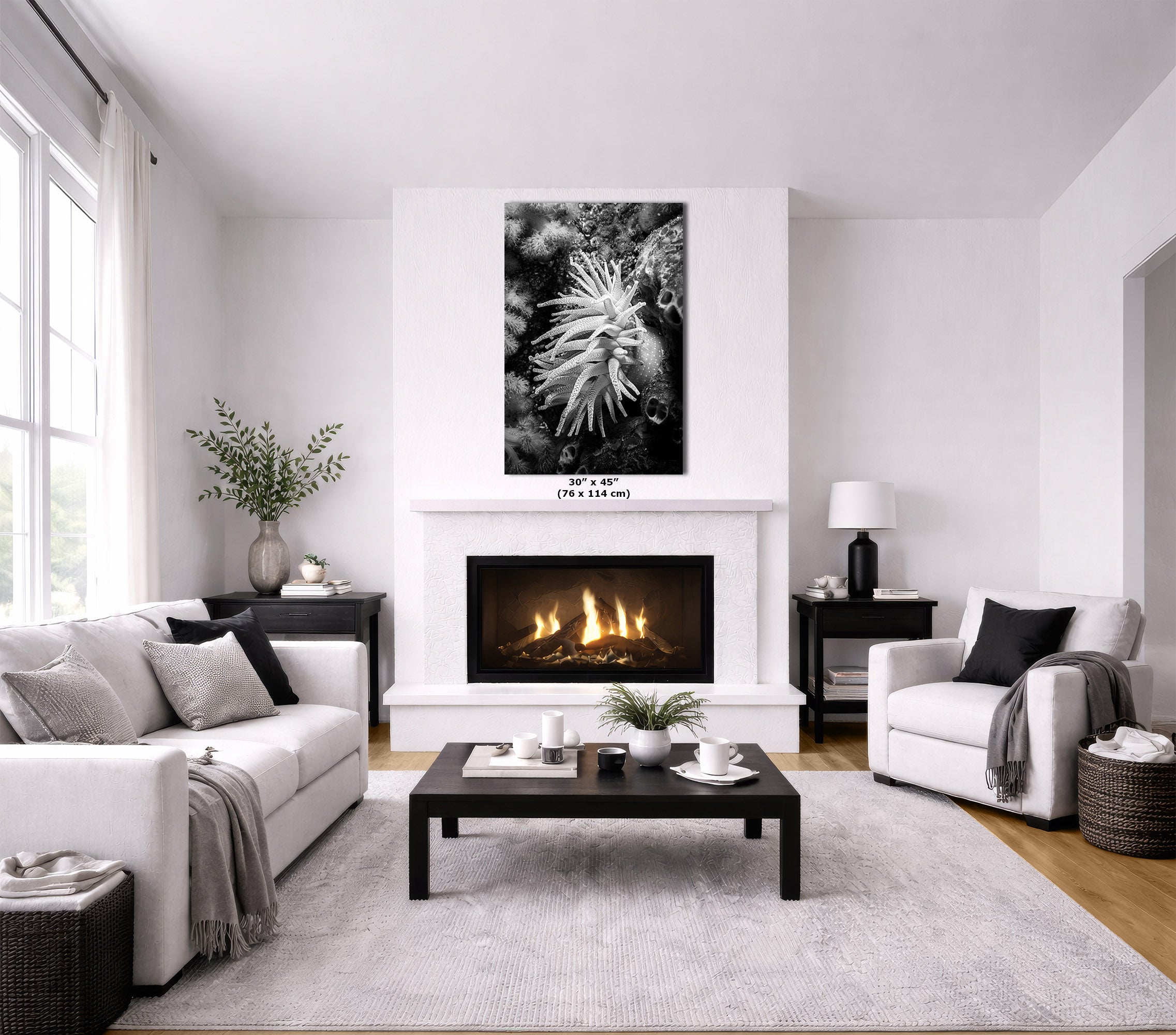 A modern living room with a white fireplace and wall, featuring a black and white floral canvas print above the fireplace. The room is styled with a white sofa, a white armchair, a dark wood coffee table, and a light gray rug.