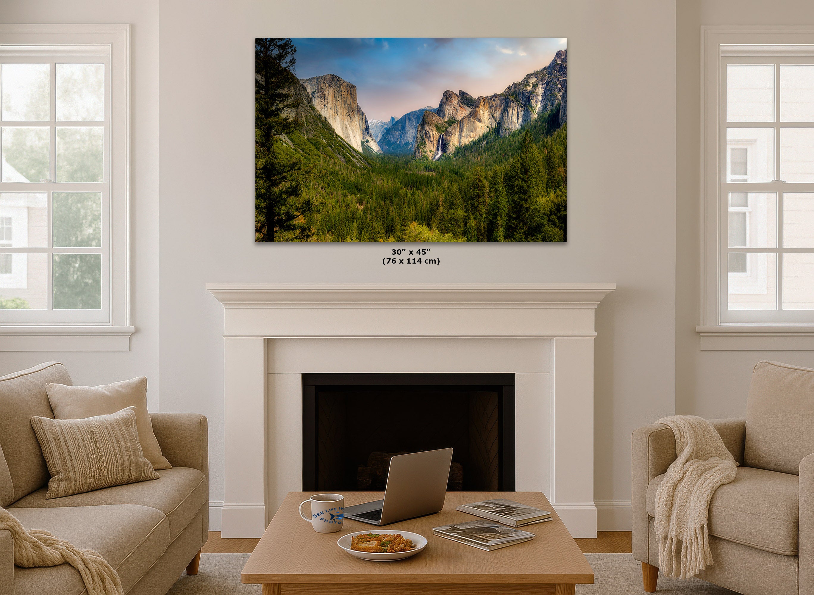 A wall art print showing a mountain valley landscape hangs above a white fireplace in a bright living room. Small text below the print reads 30 x 45 in (76 x 114 cm).