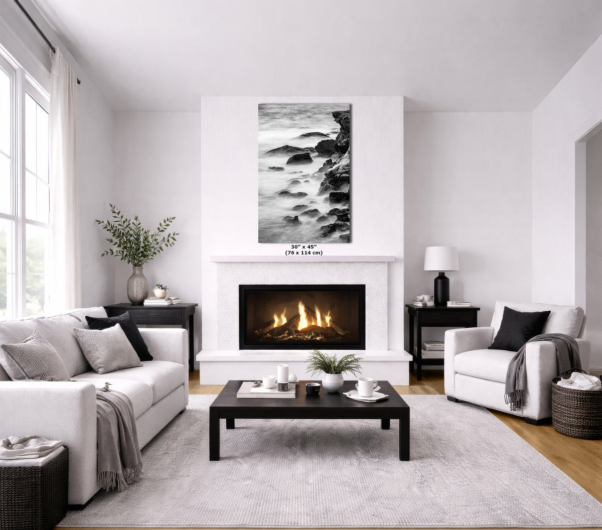 A modern living room with a white fireplace and wall, featuring a black and white landscape art print above the fireplace. The room is styled with a white sofa, a white armchair, a dark wood coffee table, and a light gray rug.