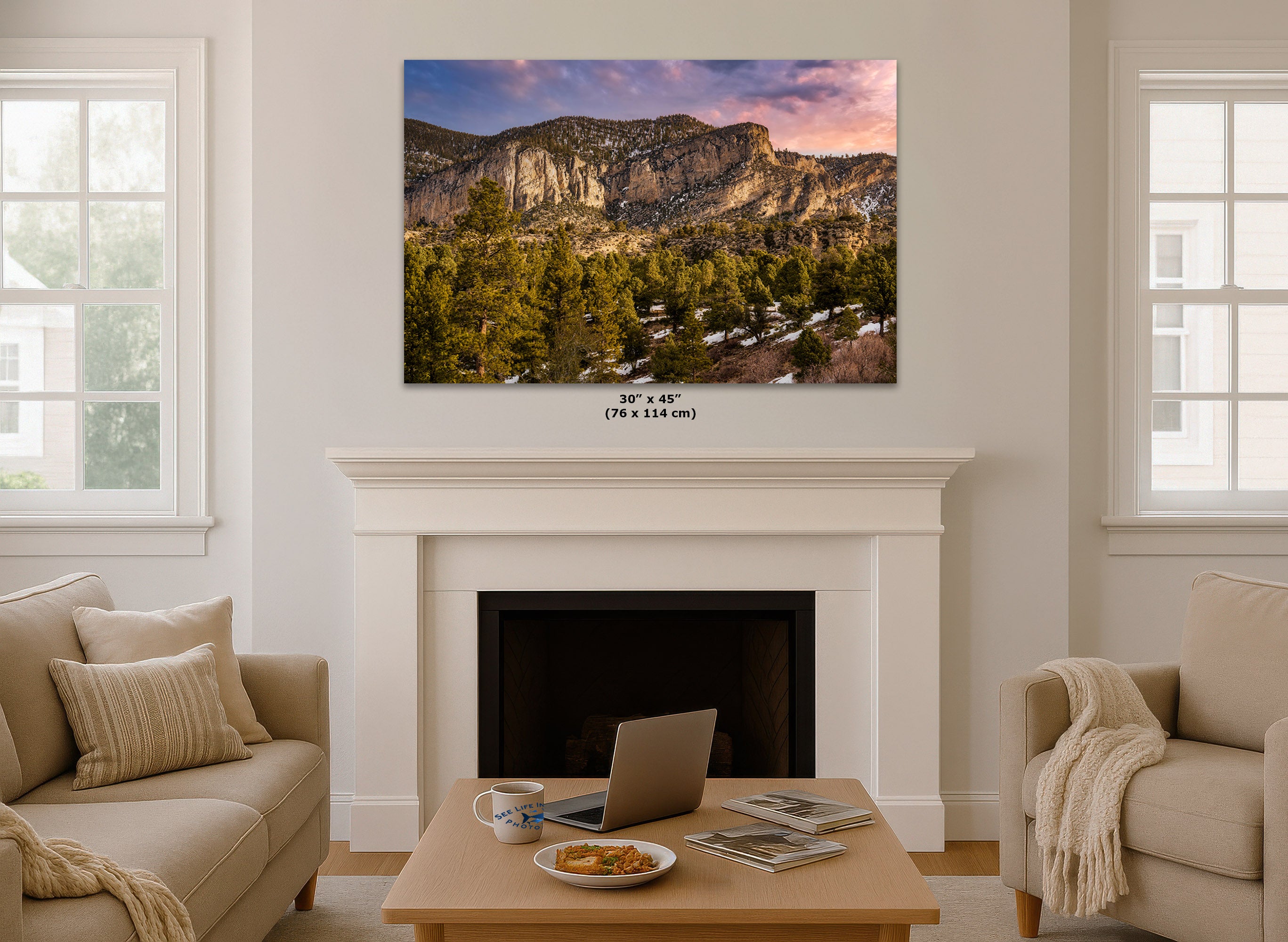 A wall art print showing a Nevada mountain landscape with evergreen trees and a pink-purple sky, displayed above a white fireplace in a bright living room. Small text below the print reads 30 x 45 in (76 x 114 cm).