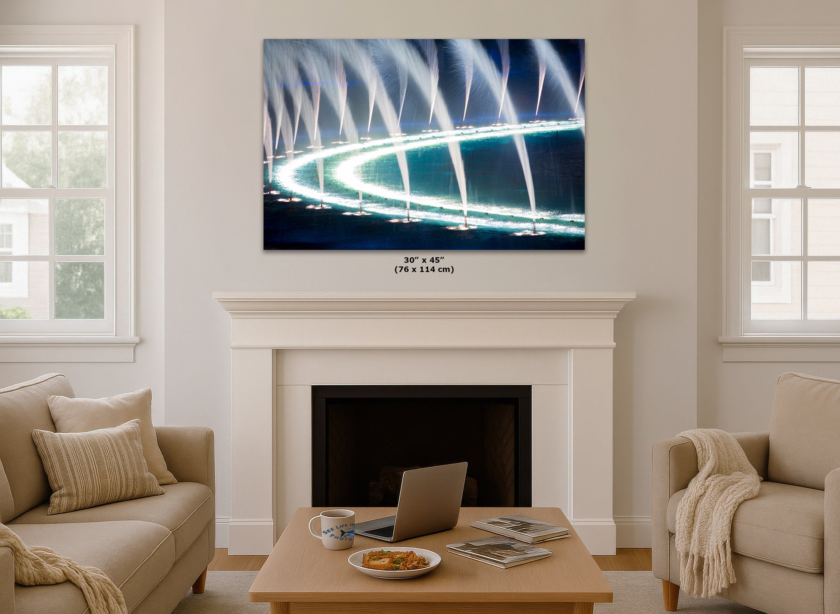 A framed wall art print showing a nighttime fountain display with bright blue and white jets, hung above a white fireplace in a styled living room with beige sofas and a coffee table.