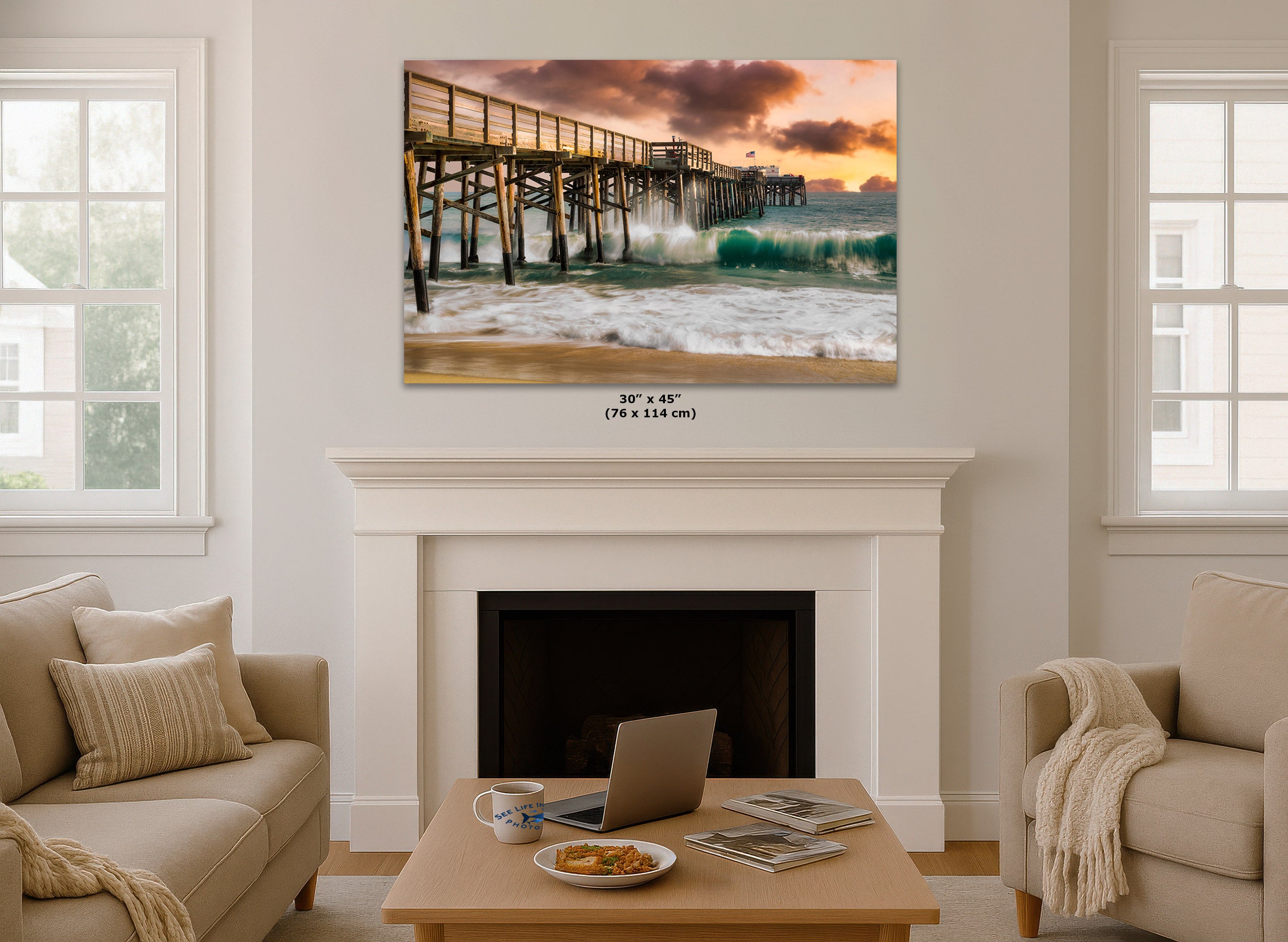 A wall art print showing a pier over ocean waves at sunset, displayed above a white fireplace in a styled living room with beige sofas and a coffee table.