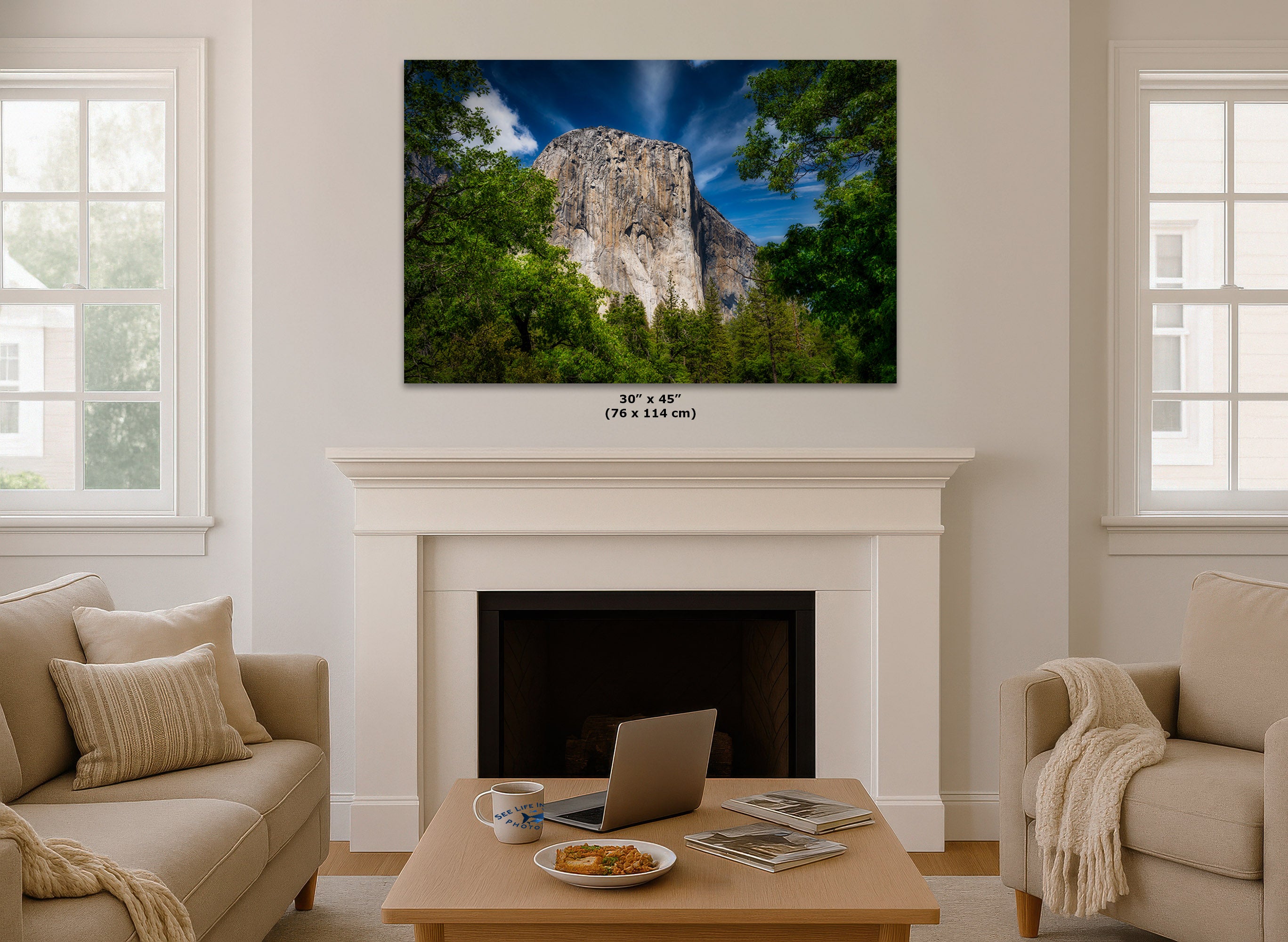 A wall art print showing a rocky mountain peak surrounded by green trees under a blue sky, displayed above a white fireplace mantel in a bright living room. Small text below the print reads 30 x 45 in (76 x 114 cm).