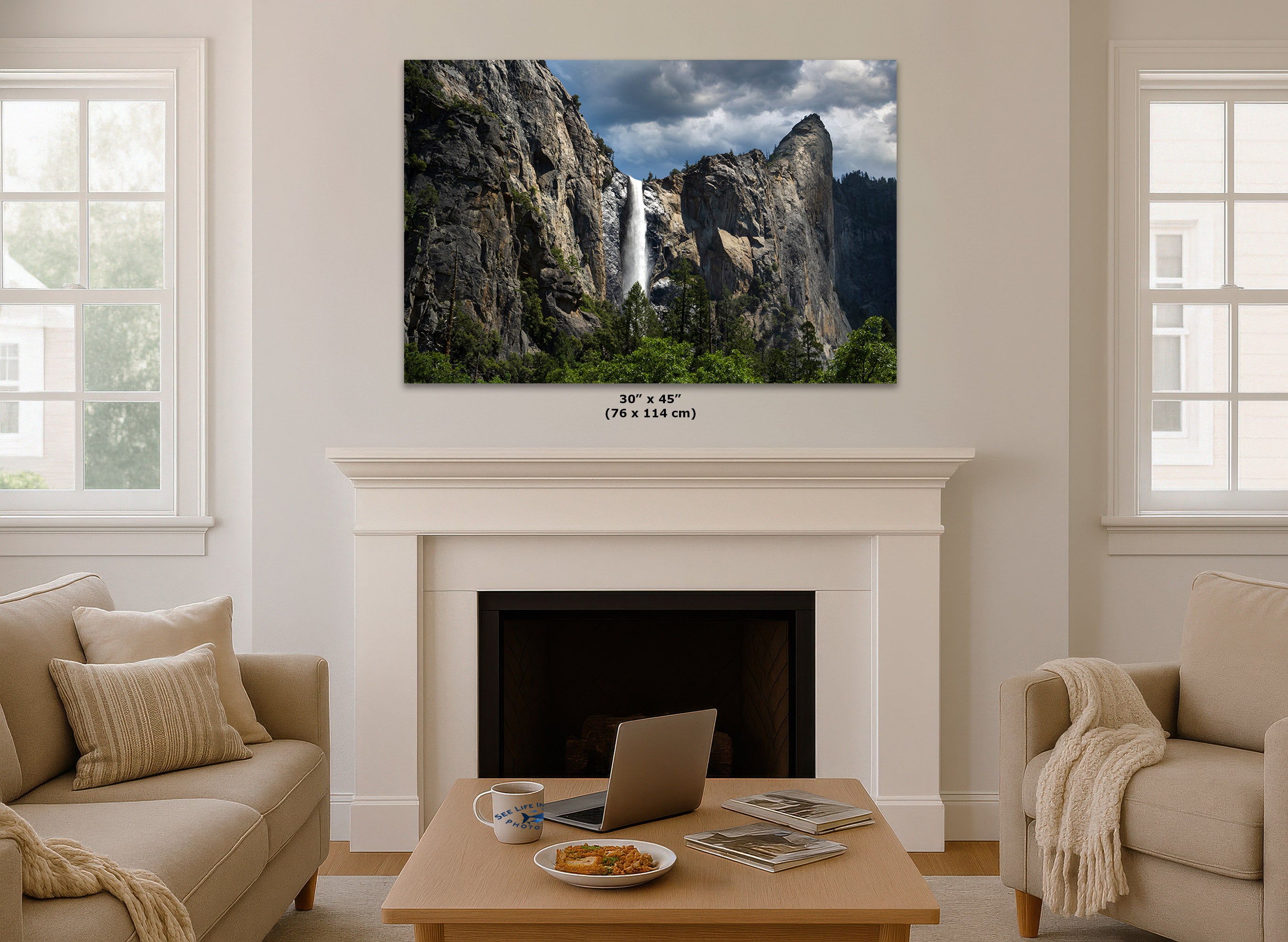 A wall art print of Yosemite Falls, shown hanging above a white fireplace mantel in a styled living room with beige sofas and a coffee table. The print includes a size label reading 30 x 45 in (76 x 114 cm).