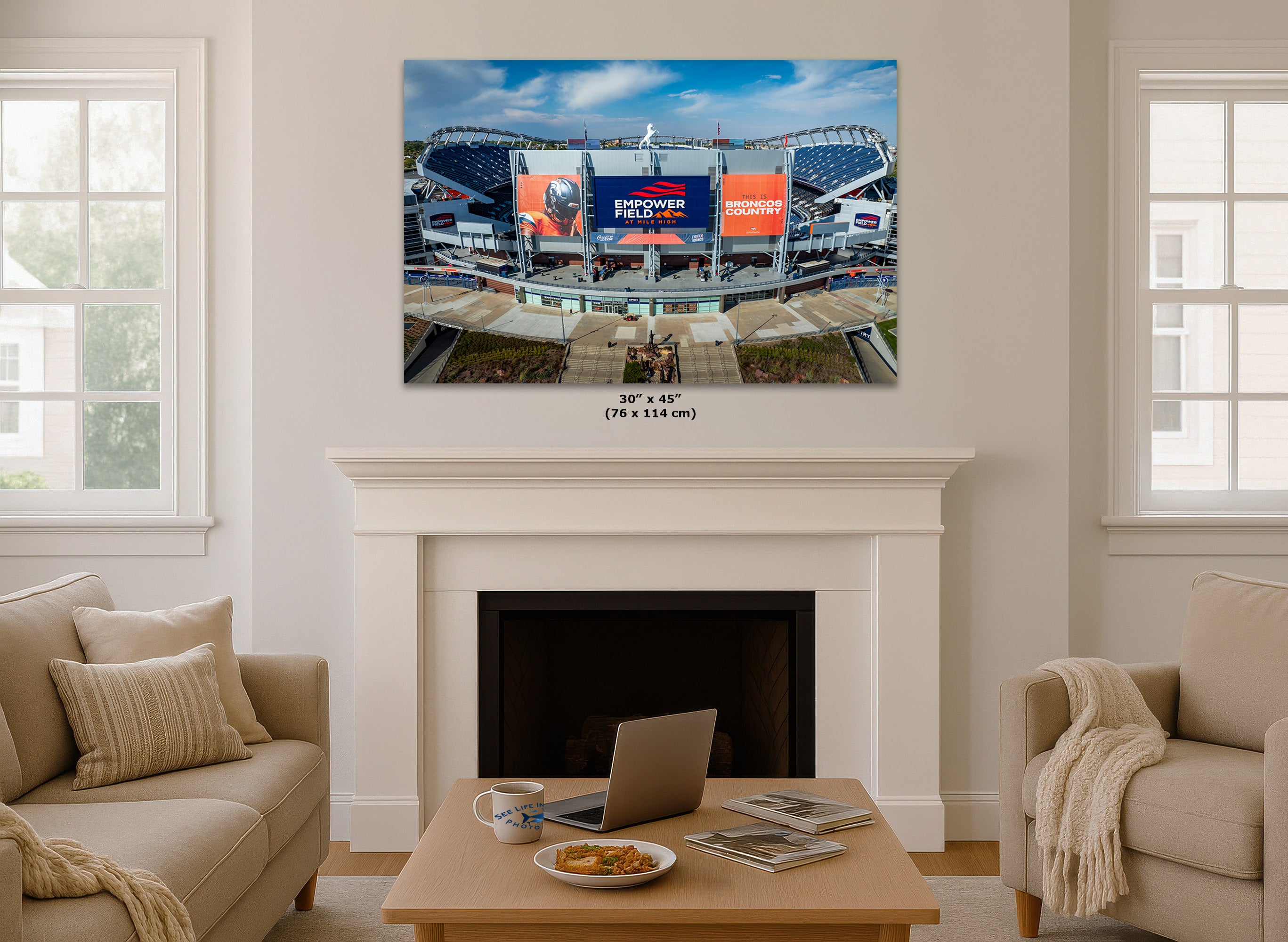 A wall art print showing a football stadium scene hangs above a white fireplace in a styled living room. The print includes visible text reading '30' x 45" (76 x 114 cm) and 'BRONCOS COUNTRY'.