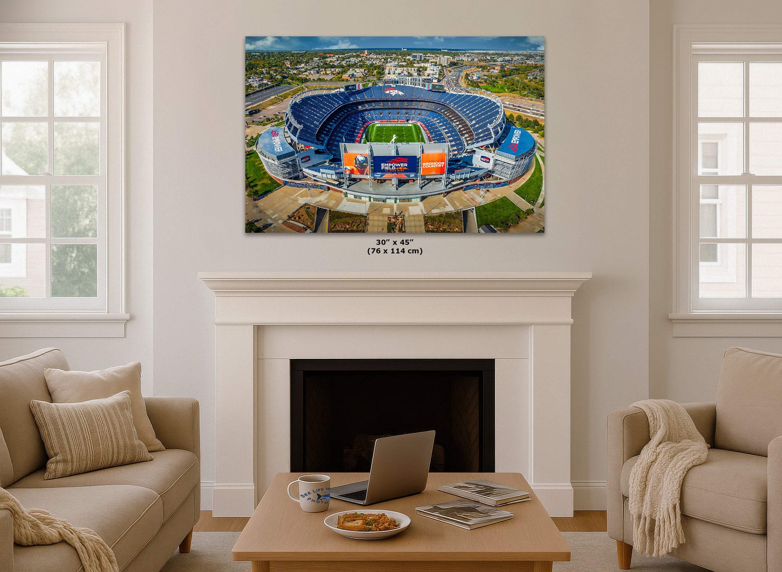 A large square wall art print showing an aerial view of a blue football stadium, displayed above a white fireplace mantel in a bright living room with beige sofas and a coffee table.