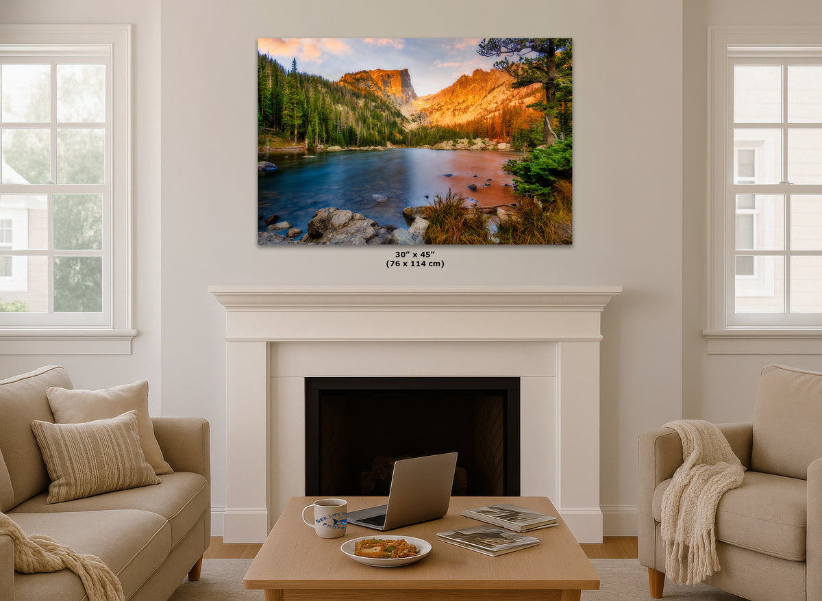 A wall art print showing a mountain lake landscape with warm sunset light, displayed above a white fireplace in a bright living room. Small text below the print reads 30 x 45 in (76 x 114 cm).