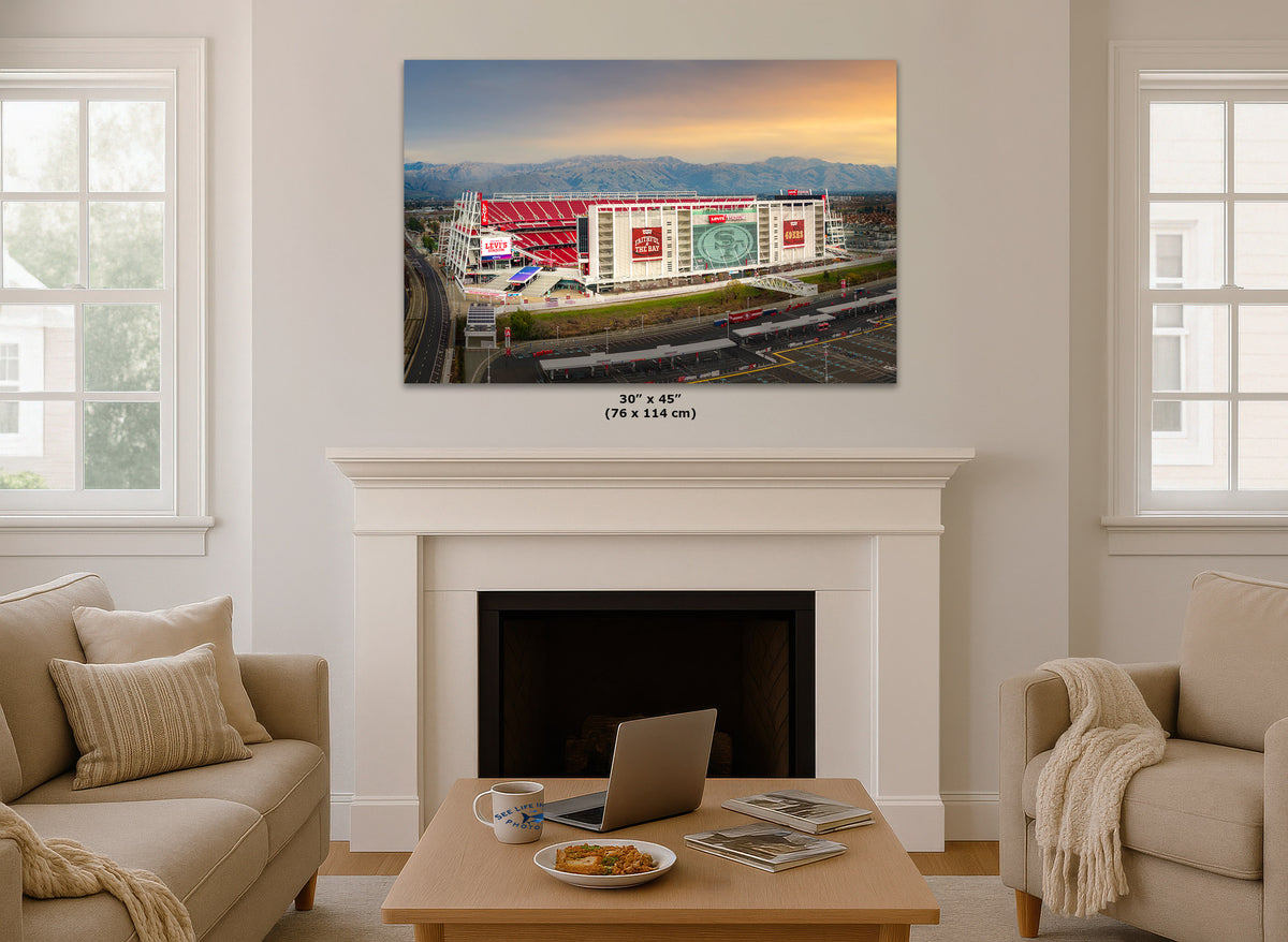 A wall art print showing an aerial view of the Levi's 49ers NFL stadium at sunset, displayed above a white fireplace mantel in a bright living room with beige sofas and a coffee table.
