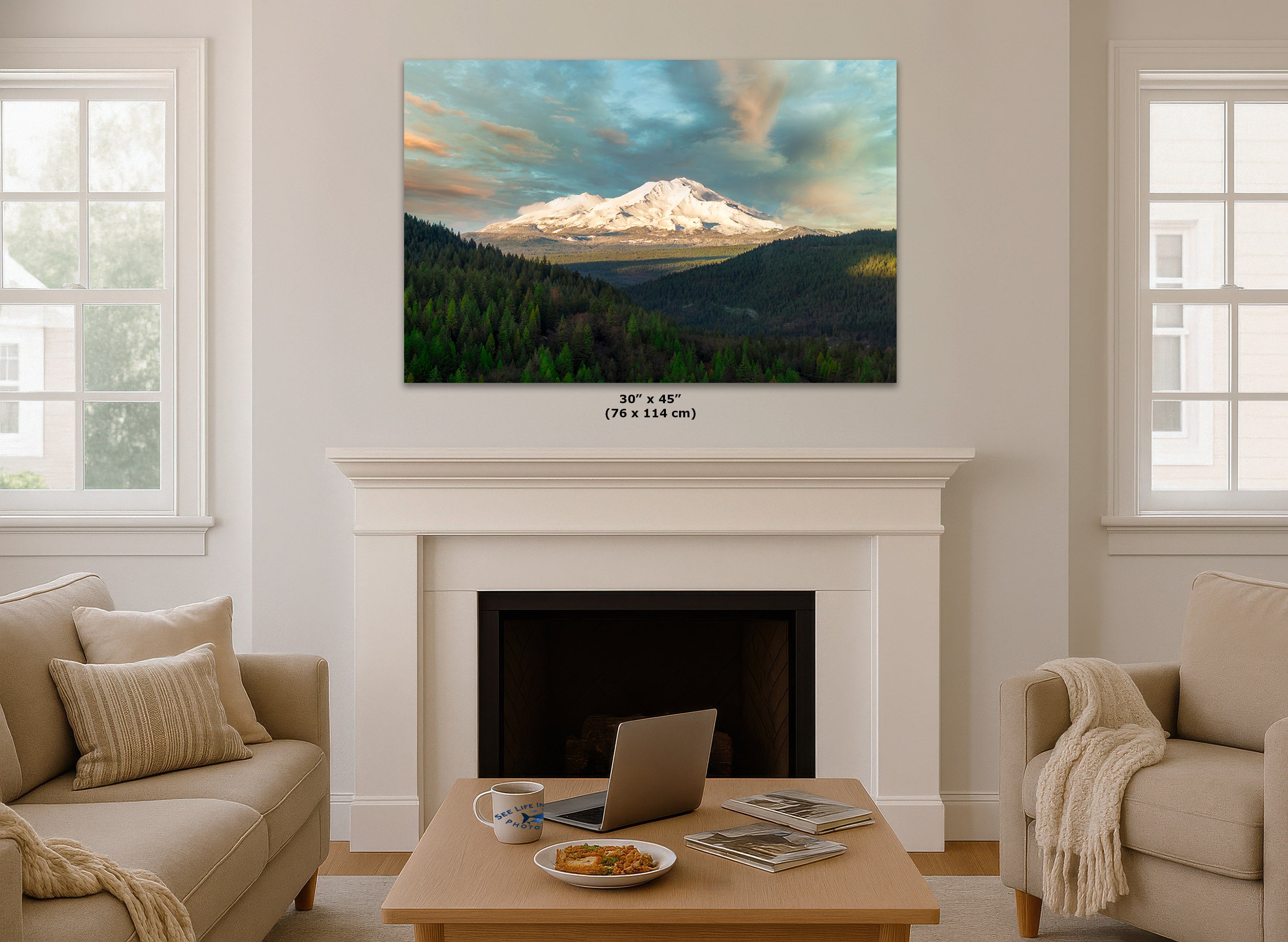 A framed wall art print of a snow-capped mountain landscape with green forests and dramatic clouds, displayed above a white fireplace mantel in a bright living room. Small text below the print reads 30 x 45 (76 x 114 cm).