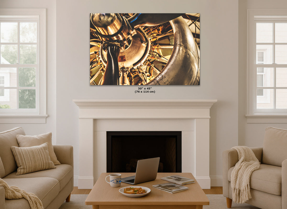 A large vintage airplane engine wall art print showing a close-up a vintage airplane engine, displayed above a white fireplace mantel in a bright living room with beige sofas and windows.
