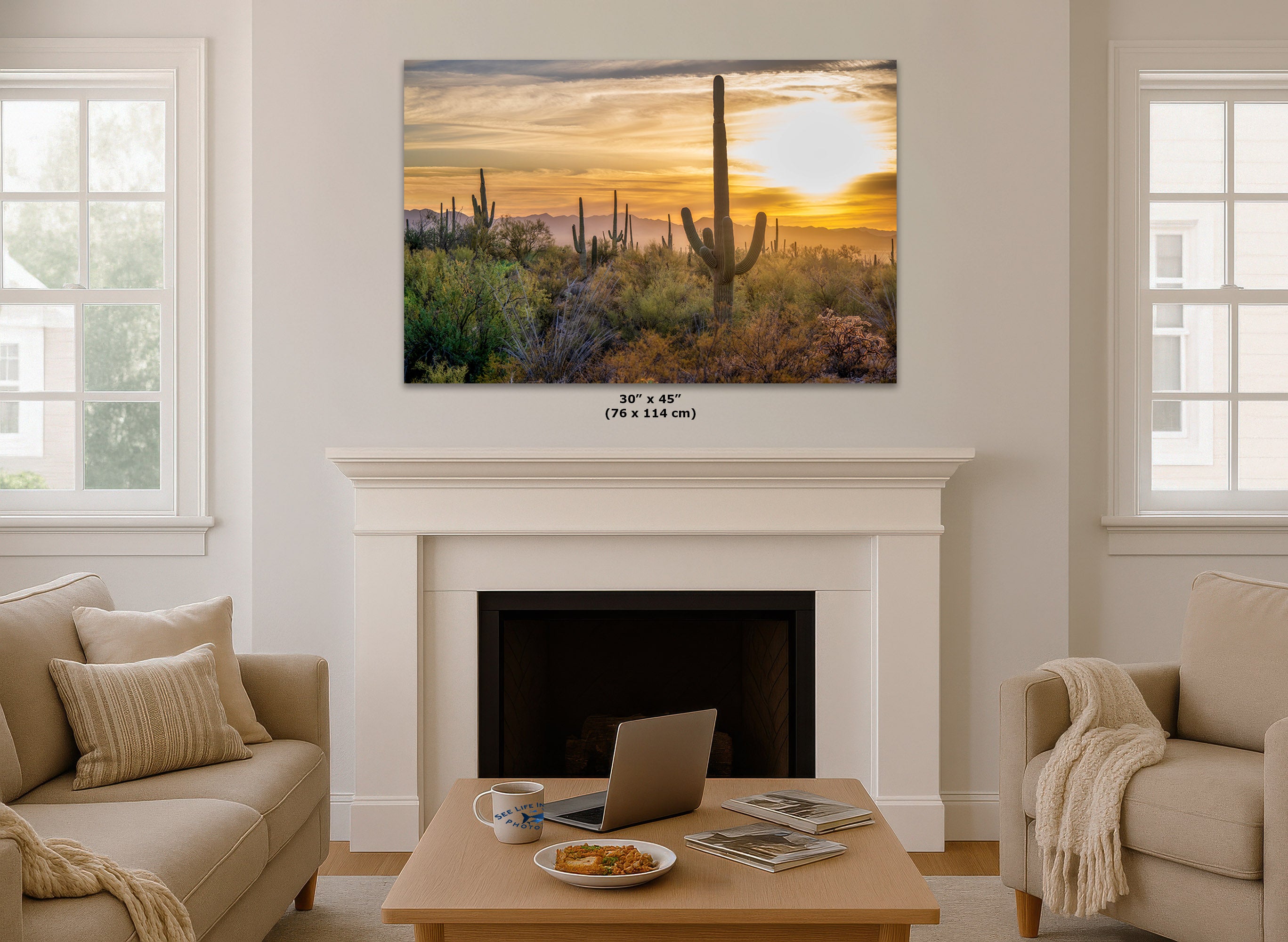 A desert landscape wall art print showing a saguaro cactus at sunset, displayed above a white fireplace mantel in a bright living room with beige sofas and a coffee table.