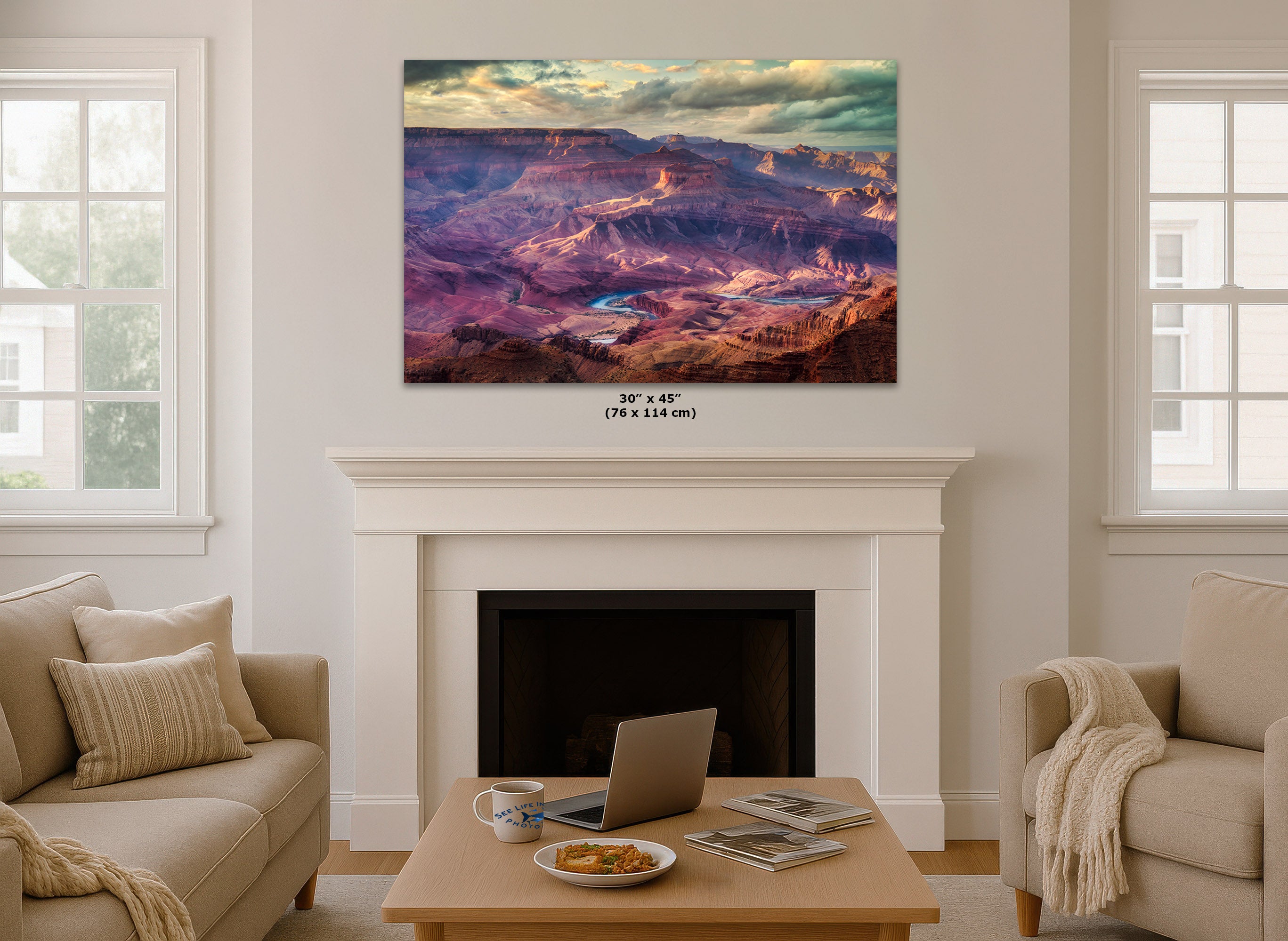 A large wall art print depicting a colorful Grand Canyon landscape is displayed above a white fireplace mantel in a bright, modern living room. The print features purple, pink, orange, and blue tones under a cloudy sky.