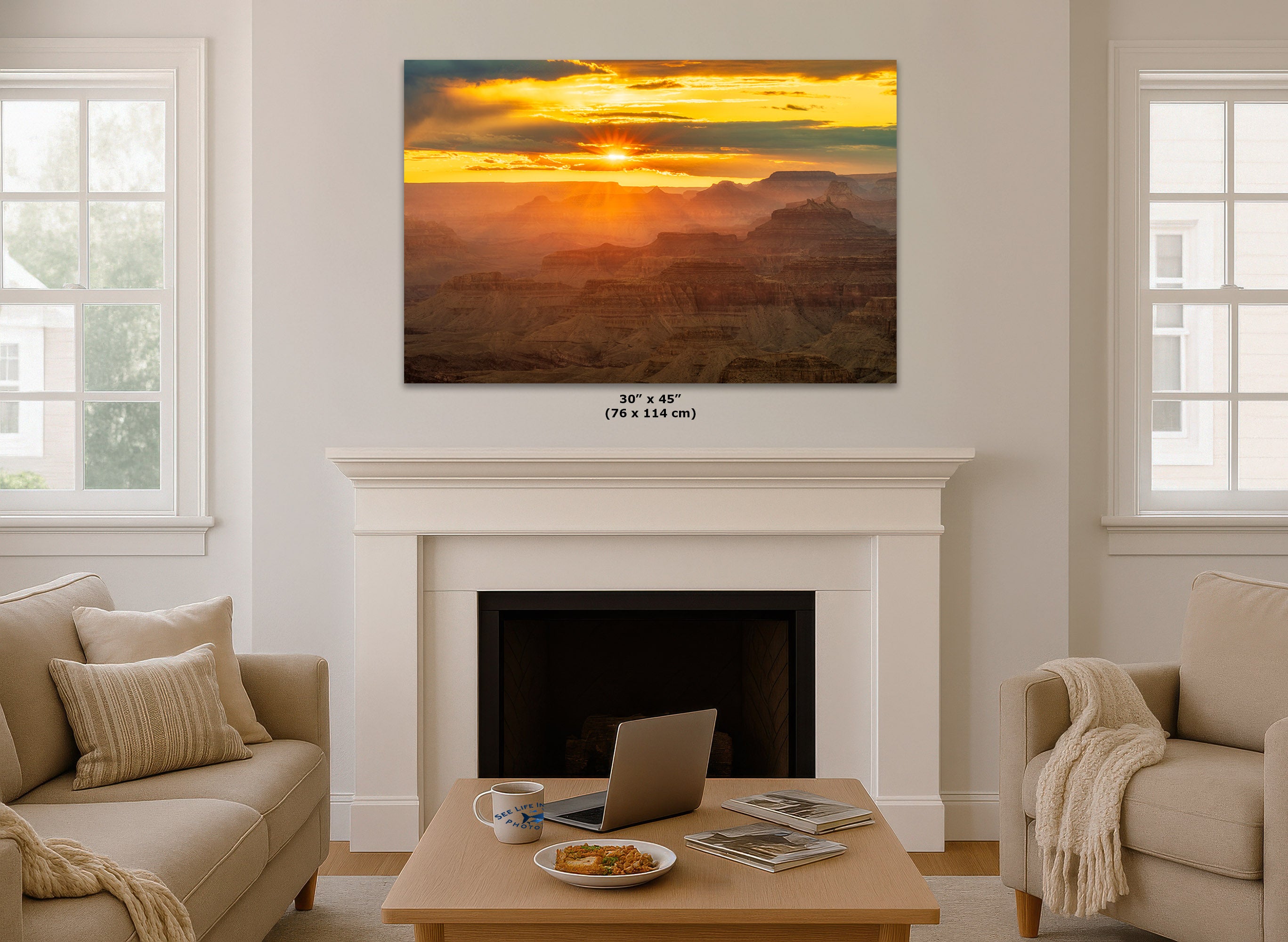 A large rectangular wall art print showing a dramatic sunset over a canyon landscape, displayed above a white fireplace mantel in a bright living room. Small text below the print reads 30 x 45 in (76 x 114 cm).