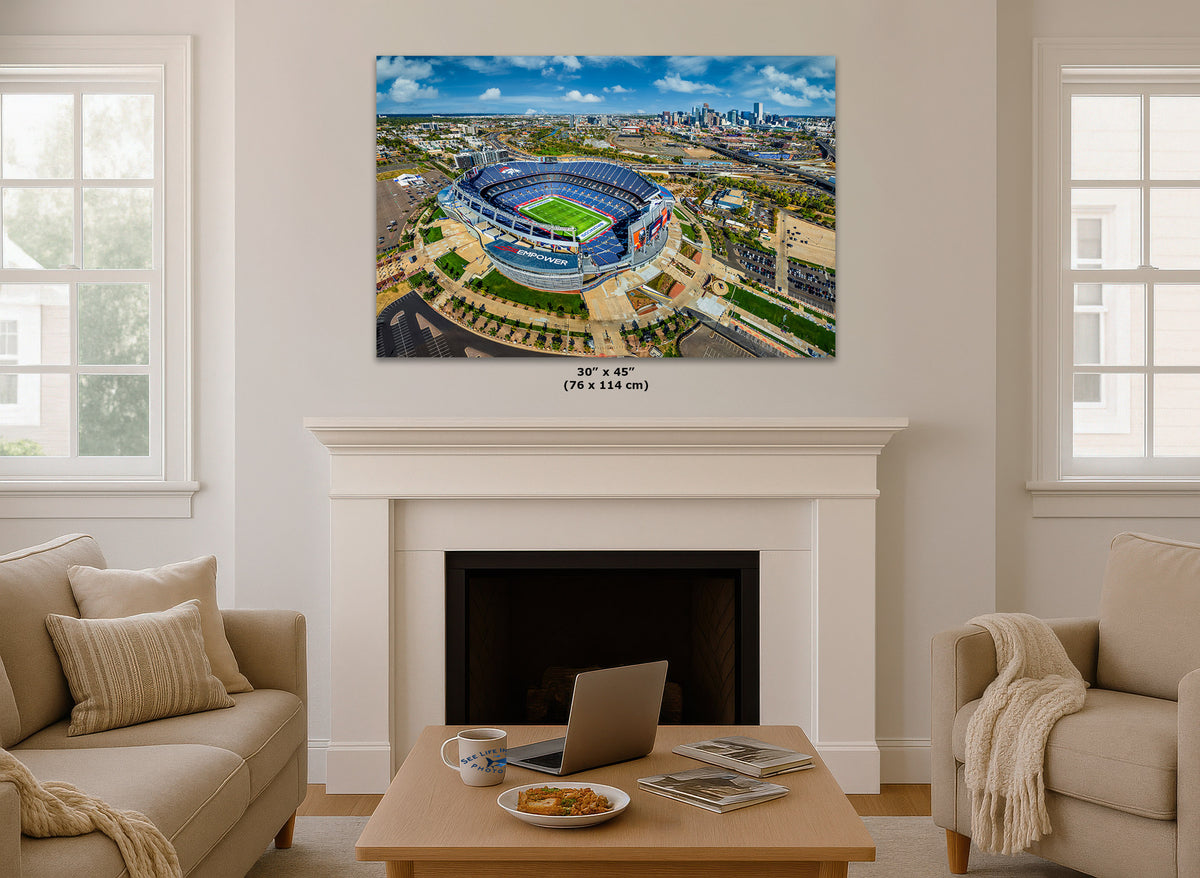 A large wall art print showing an aerial view of a stadium, displayed above a white fireplace mantel in a bright living room with beige sofas and a coffee table.
