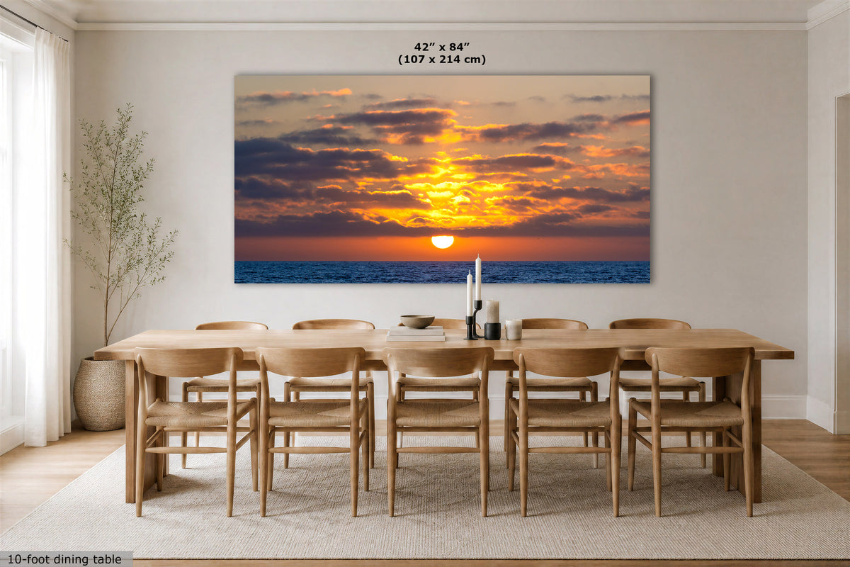 A large rectangular wall art print showing a dramatic sunset over the ocean, displayed above a wooden dining table and chairs in a bright modern dining room. The print includes size text at the top reading 42 x 84" (107 x 214 cm).