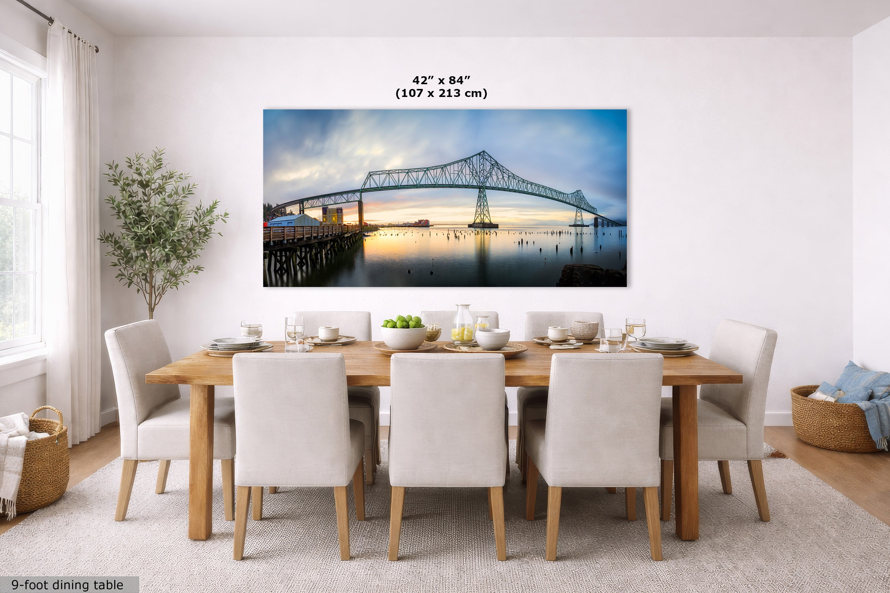 A large rectangular wall art print showing a bridge over water at dusk, displayed above a dining table in a bright modern room. The print includes size text reading 42 x 84 inches (107 x 213 cm).