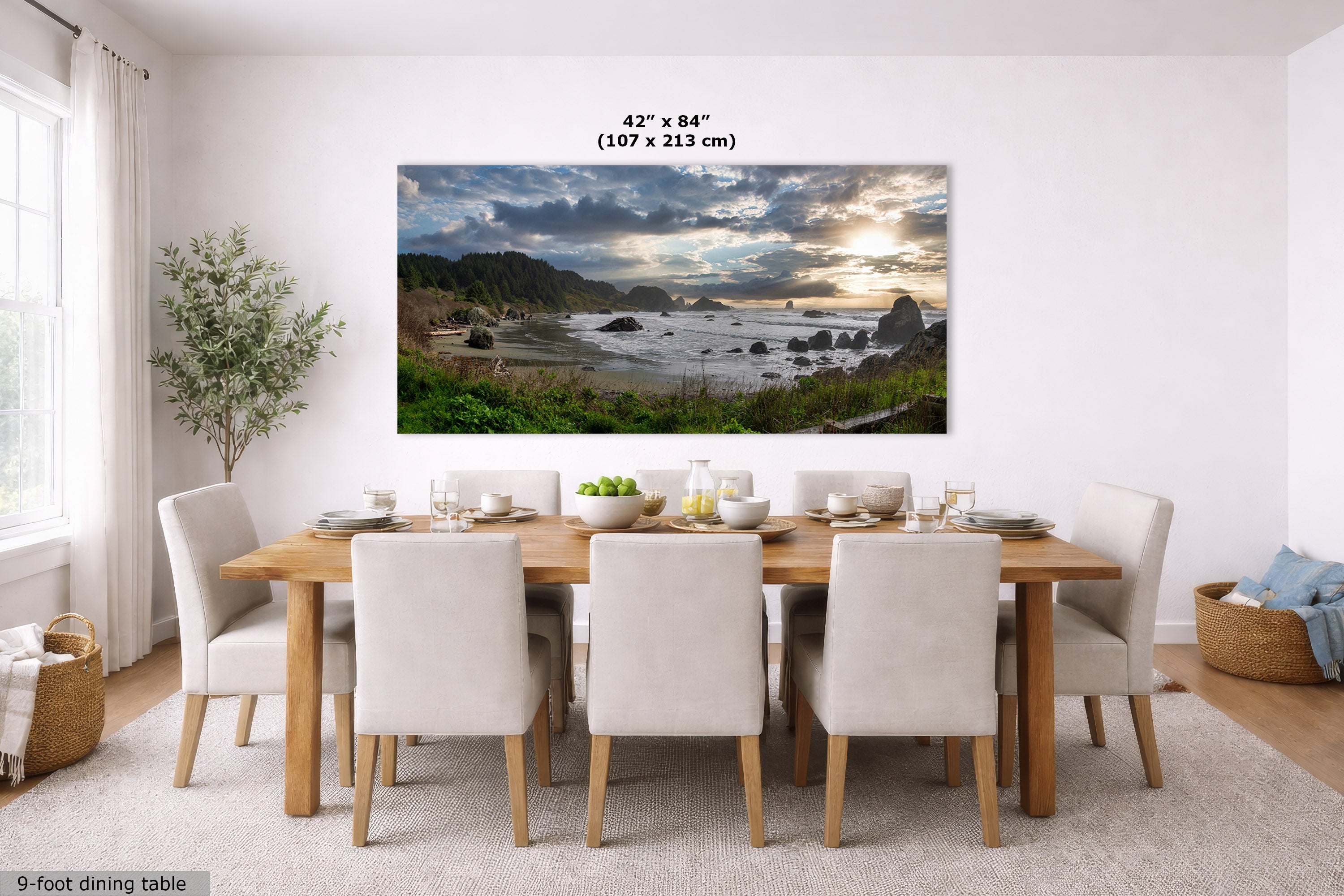 A large horizontal wall art print showing a dramatic coastal landscape with cliffs, rocks, ocean, and a glowing sunset, displayed above a dining table in a bright modern room. Text at the top of the print reads 42 x 84" (107 x 213 cm).