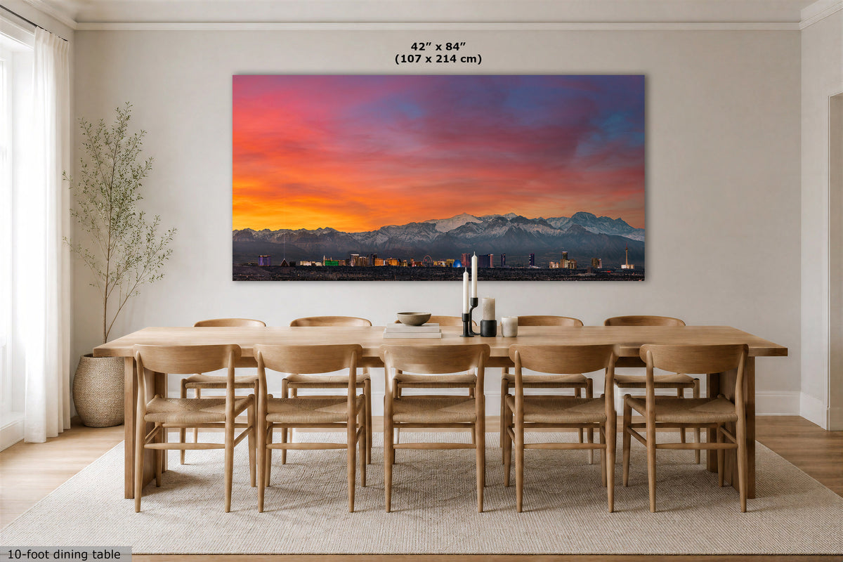 A large rectangular wall art print showing a colorful mountain sunset over a city skyline, displayed above a wooden dining table and chairs in a bright dining room. The print includes size text at the top reading 42 x 84" (107 x 214 cm).