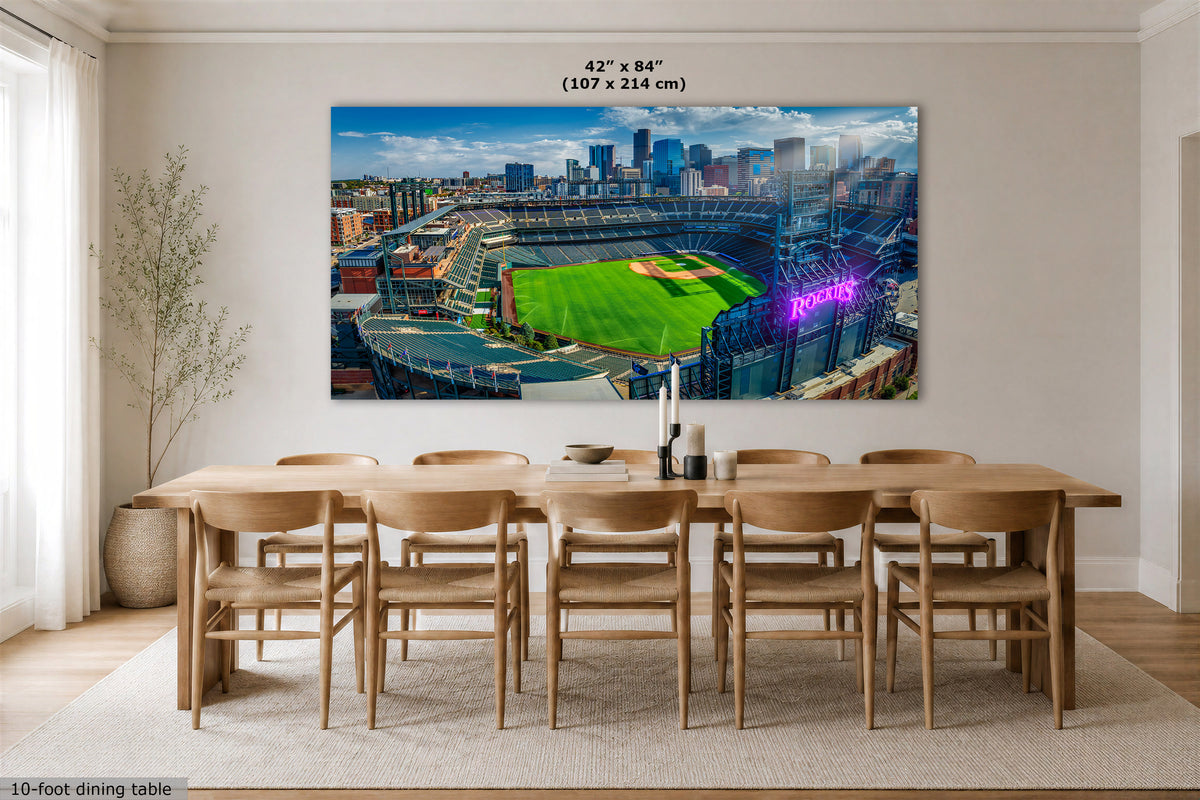 A large rectangular wall art print showing the Colorado Rockies baseball stadium in Denver with a green field, blue sky, and city skyline, displayed above a wooden dining table in a bright dining room. Text at the top reads 42 x 84" (107 x 214 cm), and text at the bottom left says 10-foot dining table.