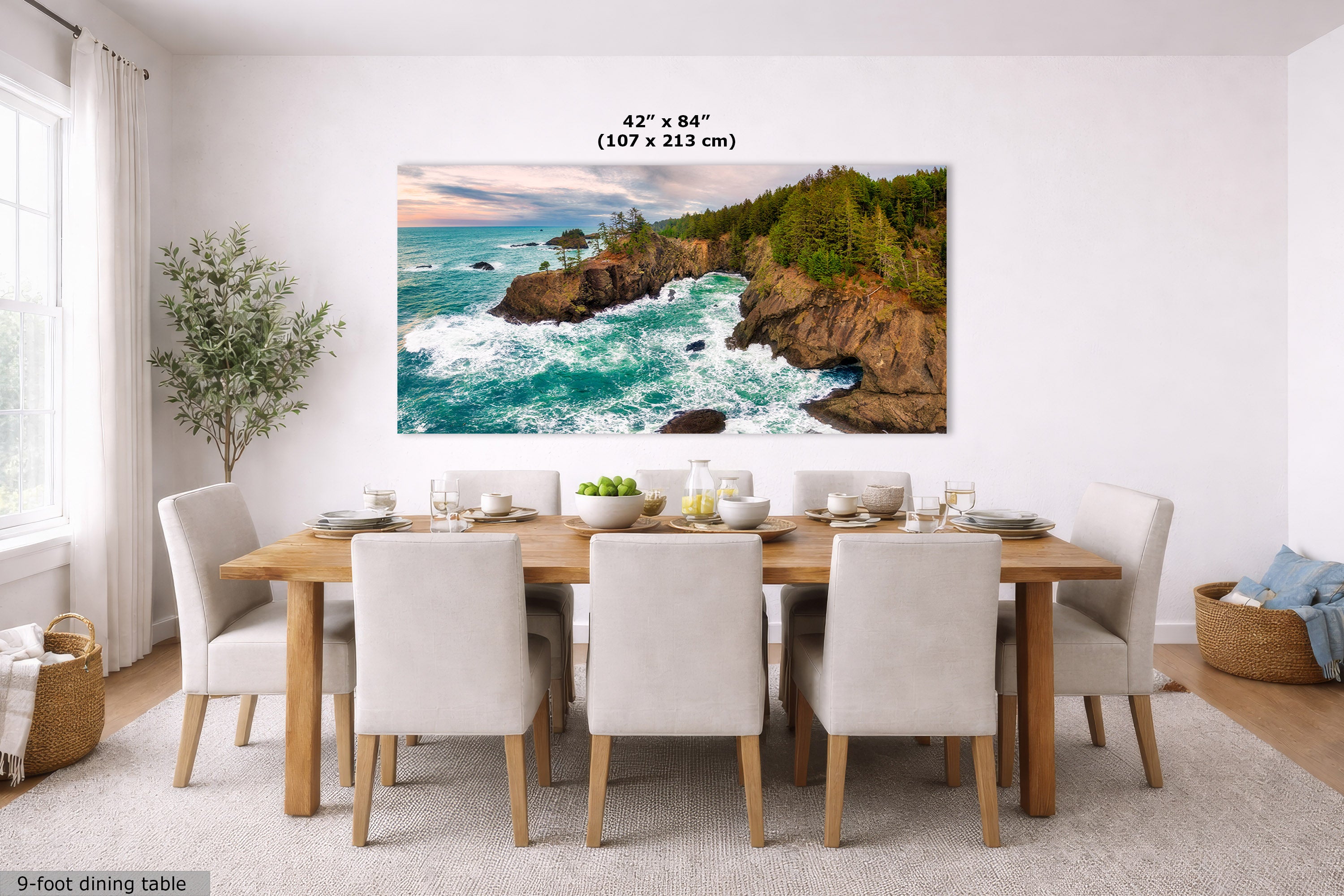 A large rectangular wall art print showing a rocky coastline with turquoise ocean waves and green cliffs, displayed above a dining table in a bright modern room. Text above the print reads 42 x 84" (107 x 213 cm).