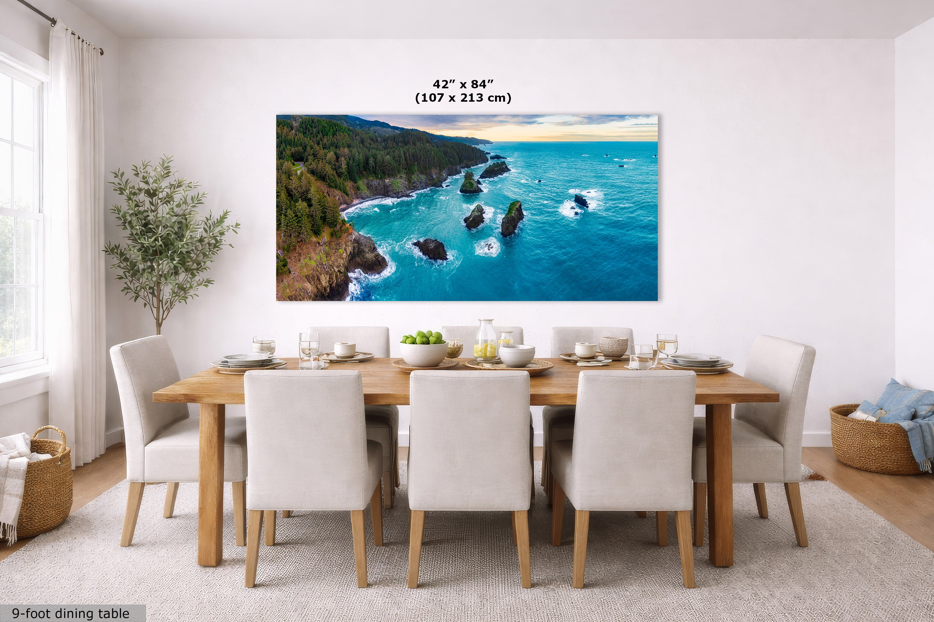A large rectangular coastal landscape wall art print hangs above a dining table in a bright modern dining room. The artwork shows turquoise ocean water, dark rocky shoreline, green forested cliffs, and a pale sky.