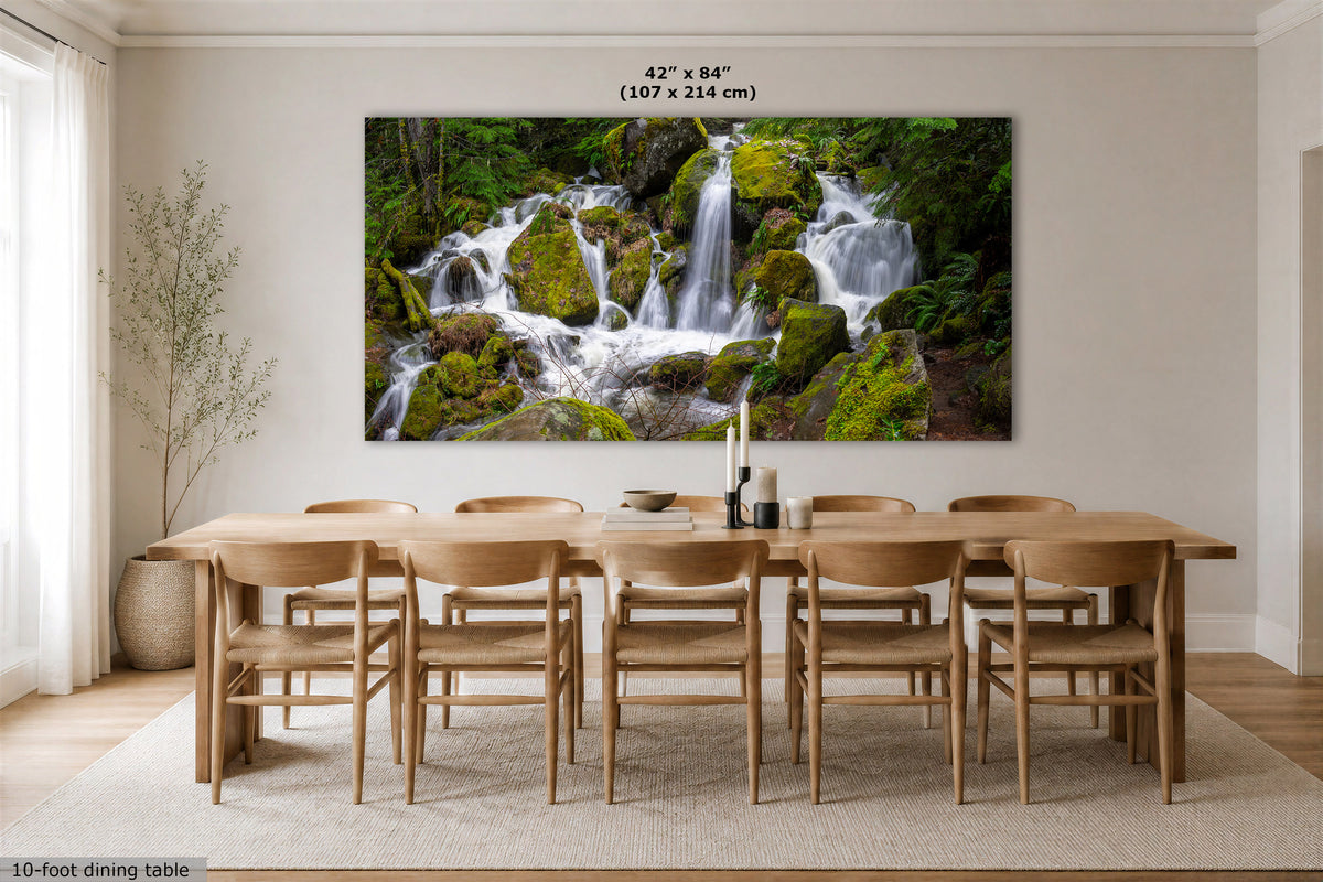 A large rectangular wall art print showing a lush green waterfall cascading over mossy rocks, displayed above a wooden dining table and chairs in a bright modern dining room. Text at the top reads 42" x 84" (107 x 214 cm).