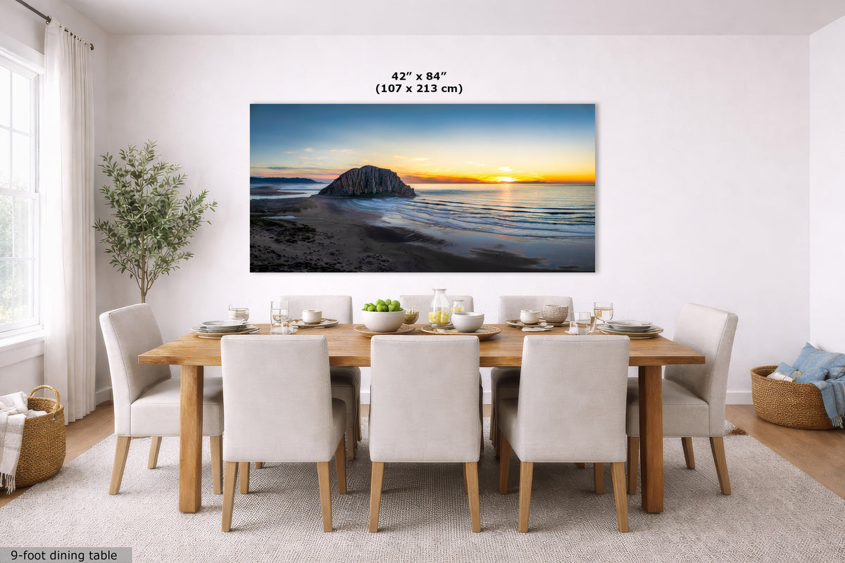 A large rectangular wall art print showing a beach and rock silhouette at sunset, displayed above a dining table in a bright modern room. Text above the print reads 42 x 84 inches (107 x 213 cm).