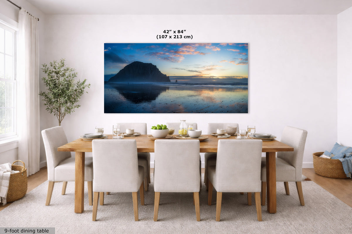 A large panoramic wall art print showing a beach and ocean sunset with a mountain silhouette, displayed above a dining table in a bright modern room. The print includes size text reading 42 x 84 inches (107 x 213 cm).