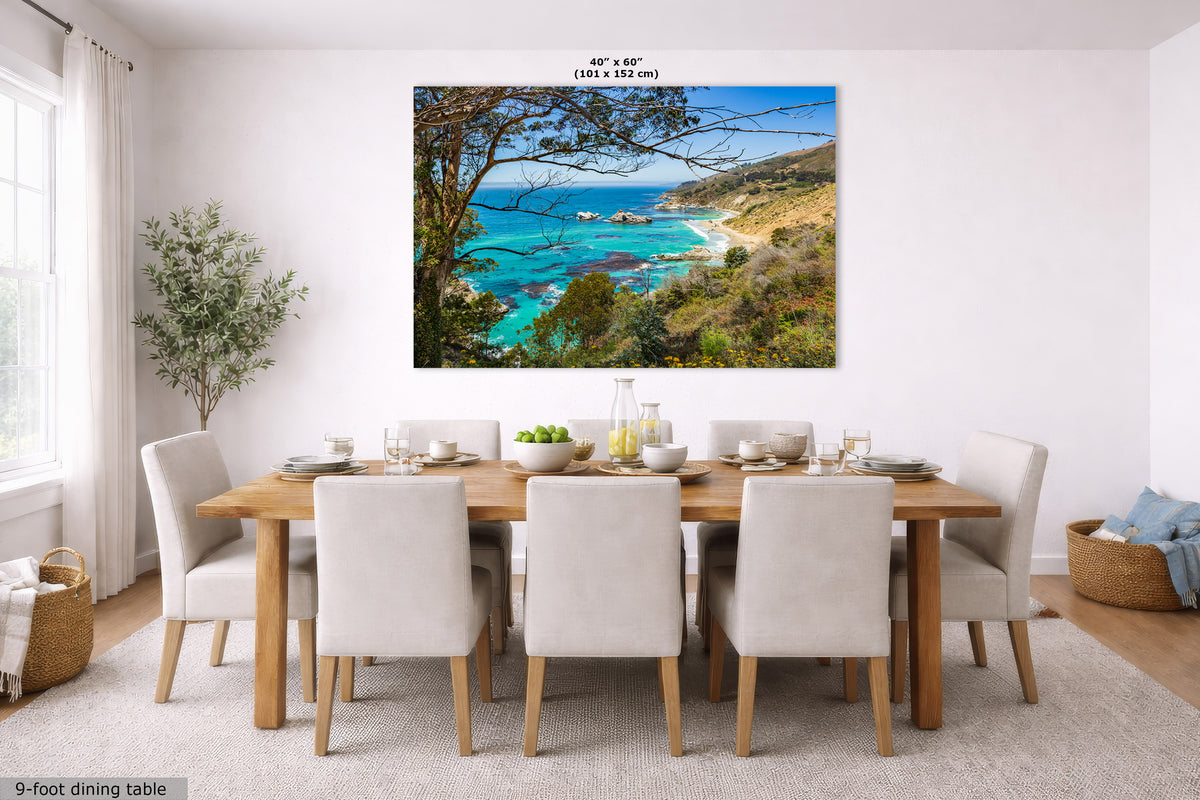 A large wall art print showing a coastal landscape with turquoise ocean, sandy beach, and green hills, displayed above a wooden dining table with white chairs in a bright dining room. Text at the top of the print reads 40" x 60".