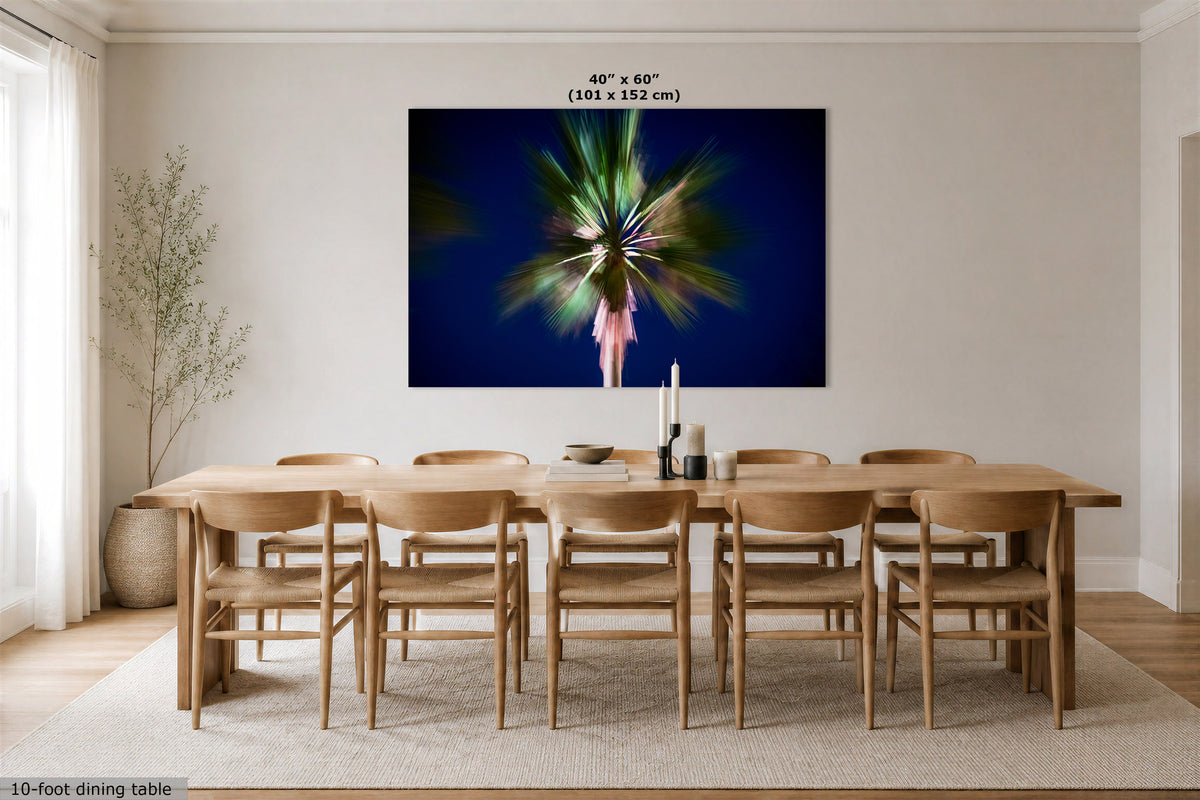 A large wall art print hangs above a wooden dining table and chairs in a bright, modern dining room. The artwork shows a stylized green palm tree against a deep navy blue background with a blurred figure in pink.