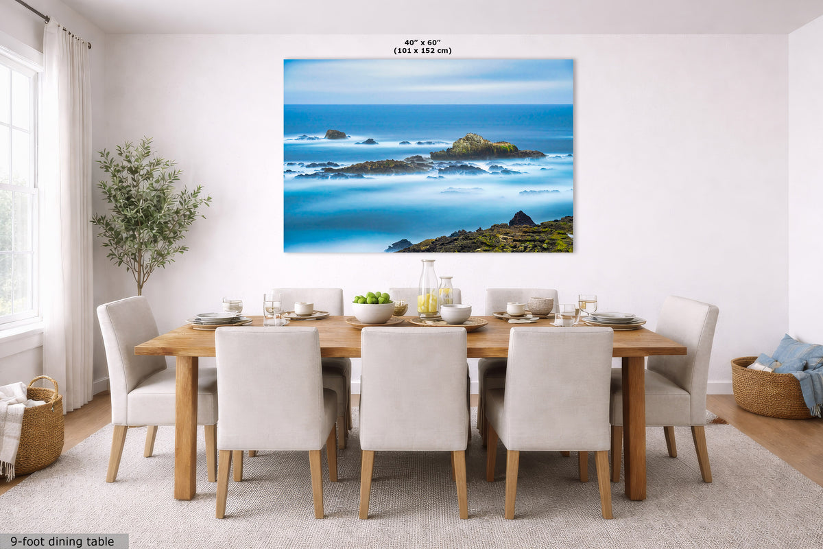 A large wall art print showing a coastal ocean scene with blue water, rocks, and green shoreline, displayed above a dining table in a bright modern room. Small text at the top reads 40 in x 60 in (101 x 152 cm), and text at the bottom left says 9-foot dining table.