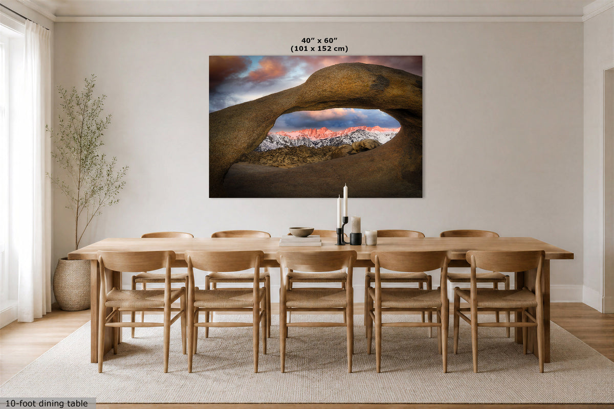 A large wall art print showing a natural stone arch over a mountain landscape at sunset, displayed above a wooden dining table and chairs in a bright dining room. The print includes the text '40" x 60" (101 x 152 cm)' at the top.