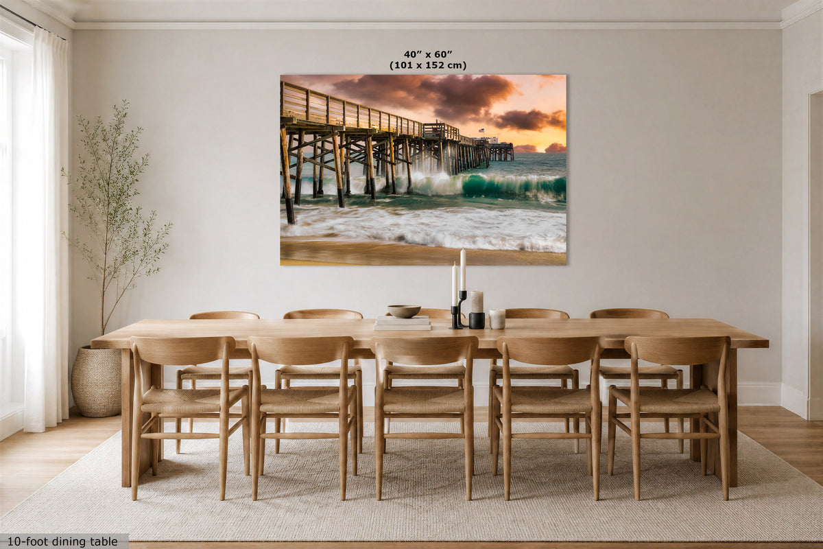 A large rectangular wall art print showing a pier over ocean waves at sunset, displayed above a wooden dining table and chairs in a bright dining room. Text at the top of the print reads 40" x 60" (101 x 152 cm).