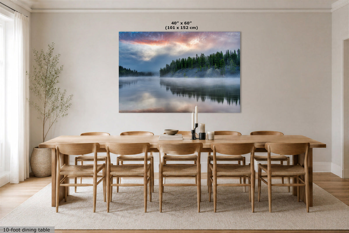 A 40" x 60" canvas print of a misty forest lake at sunrise, displayed on a light beige wall above a wooden dining table and chairs in a modern dining room.