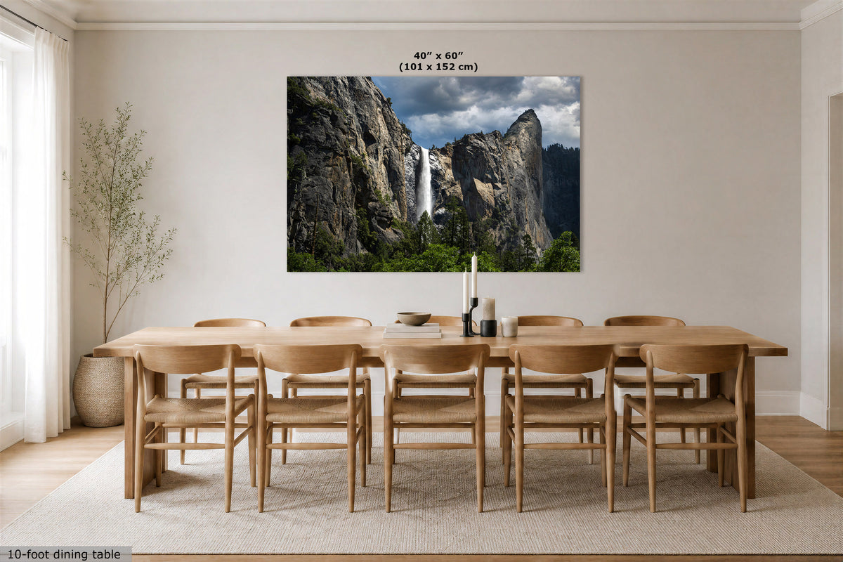 A large wall art print depicting a waterfall between rocky cliffs, shown hanging above a wooden dining table and chairs in a bright, modern dining room. The print includes size text at the top reading 40" x 60" (102 x 152 cm).