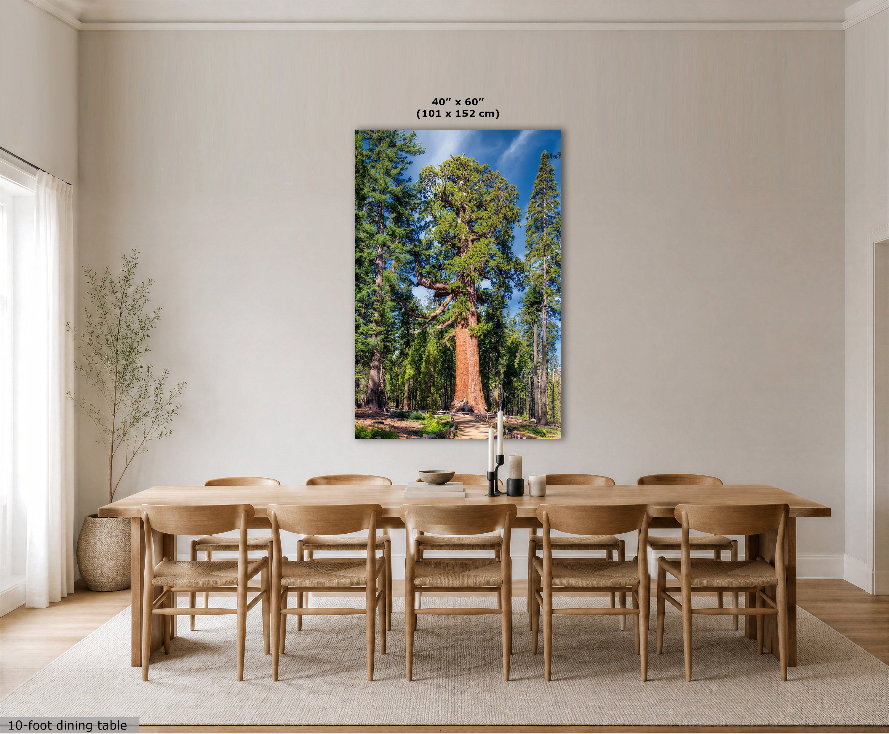 A large vertical wall art print showing a forest scene with a large tree under a blue sky, displayed above a wooden dining table and chairs in a bright dining room. The print includes size text at the top and a small text label at the bottom left.