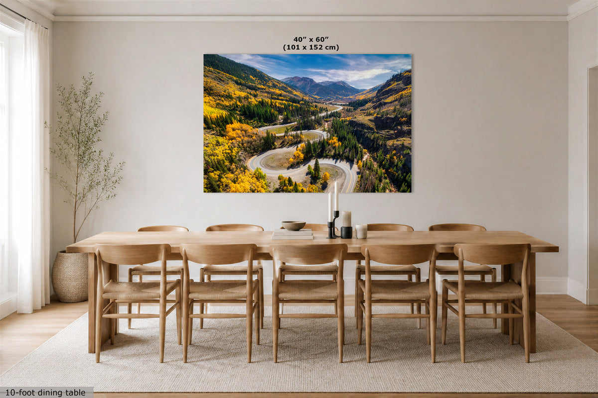 A large rectangular wall art print showing a mountain valley with autumn trees and a winding road, displayed above a wooden dining table and chairs in a bright dining room. Text at the top reads 40" x 60" (101 x 152 cm).