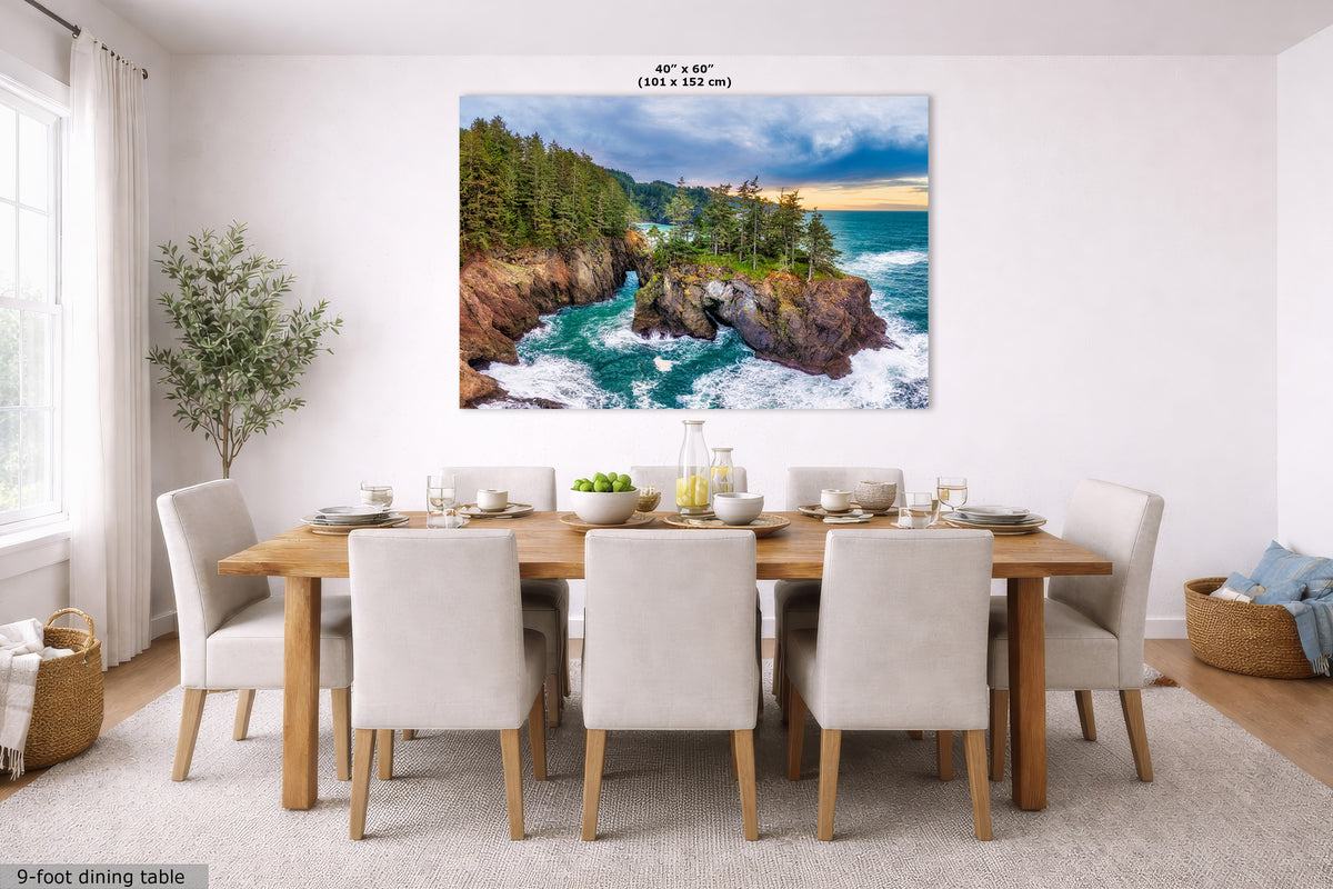 A large square wall art print showing a rocky coastal ocean scene with green trees and a blue sky, displayed above a dining table in a bright modern room. The print includes size text at the top and a small label at bottom left.
