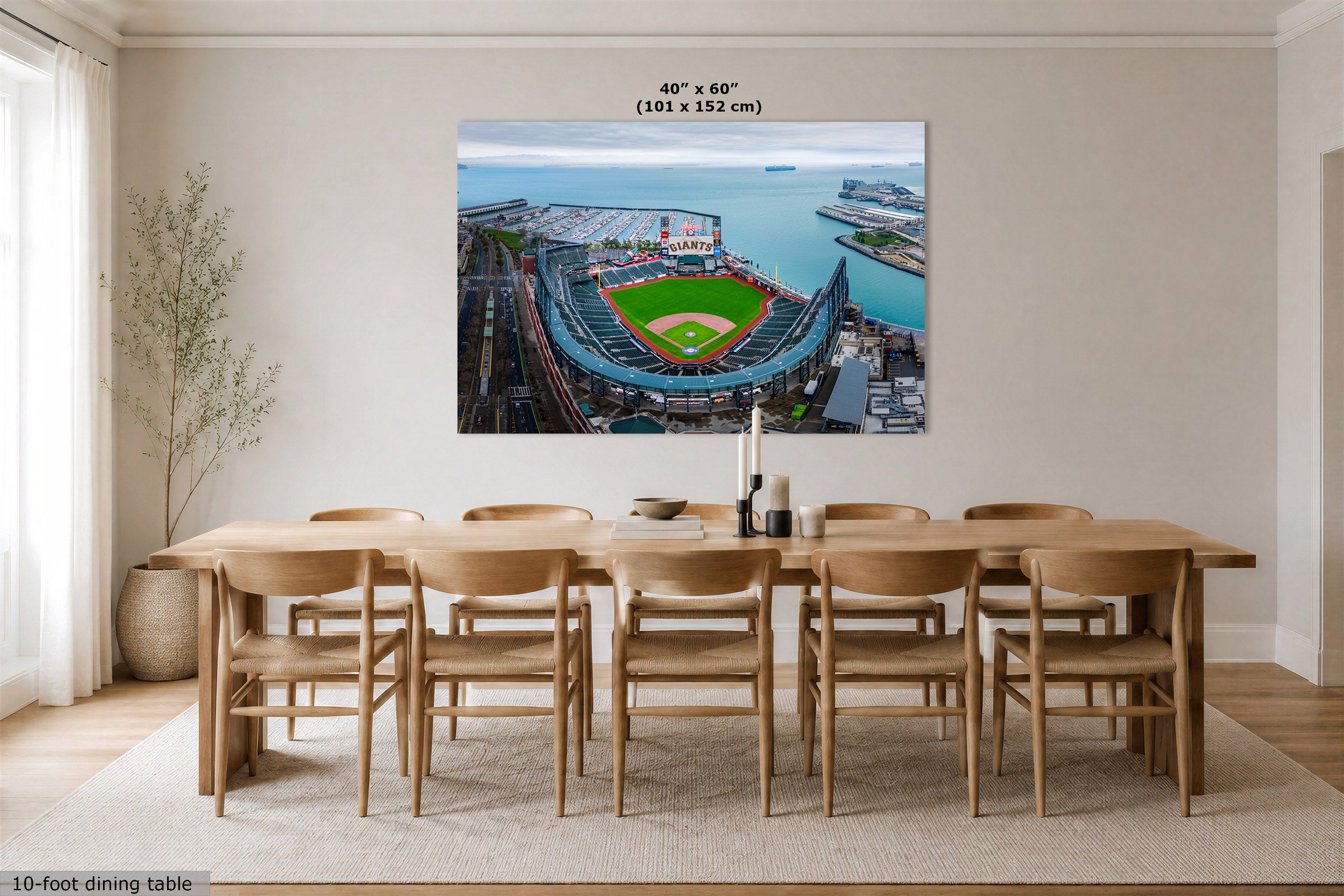 A large wall art print showing an aerial view of a baseball stadium by the water, displayed above a wooden dining table and chairs in a bright modern room. The print includes size text at the top reading 40" x 60" (101 x 152 cm).
