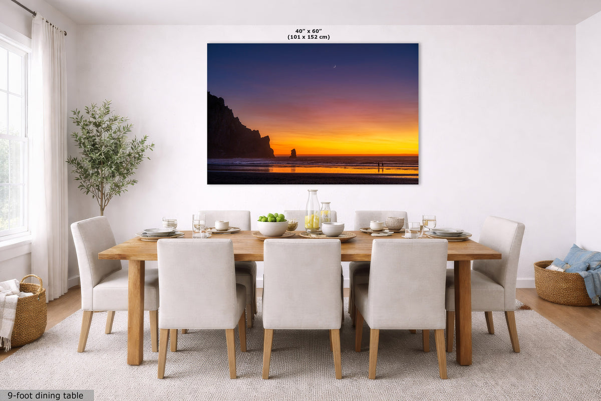 A large rectangular wall art print showing a sunset beach scene with cliffs and ocean, displayed above a dining table in a bright modern room. The print includes size text at the top and a small label at bottom left.