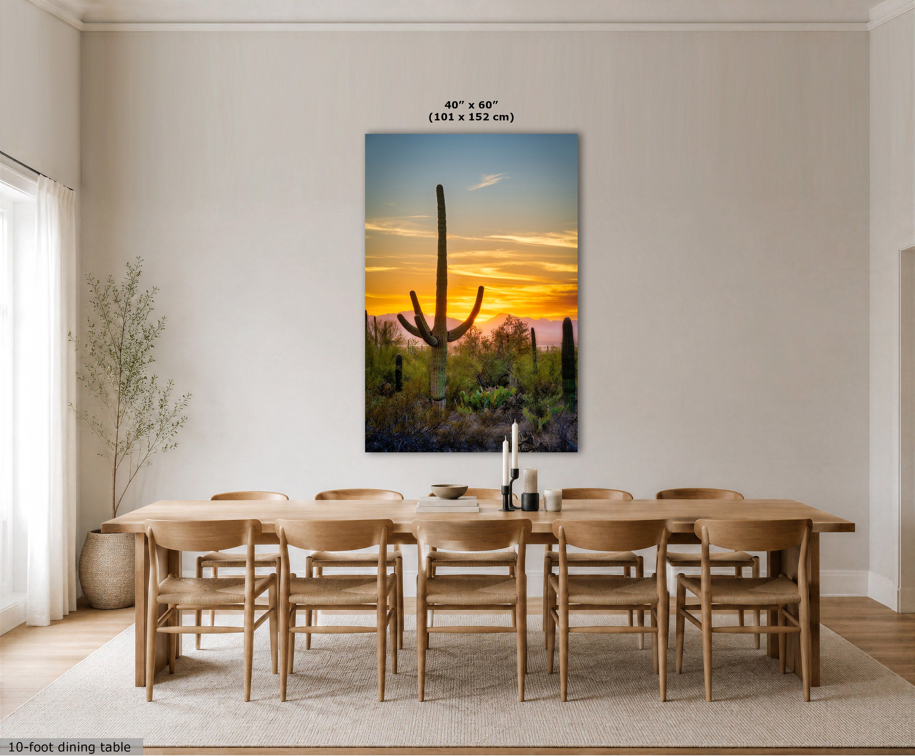 A vertical canvas wall art print depicting a desert sunset with saguaro cacti, displayed in a modern dining room above a wooden dining table and chairs. The print features a warm orange and yellow sunset sky with blue and green desert tones below.